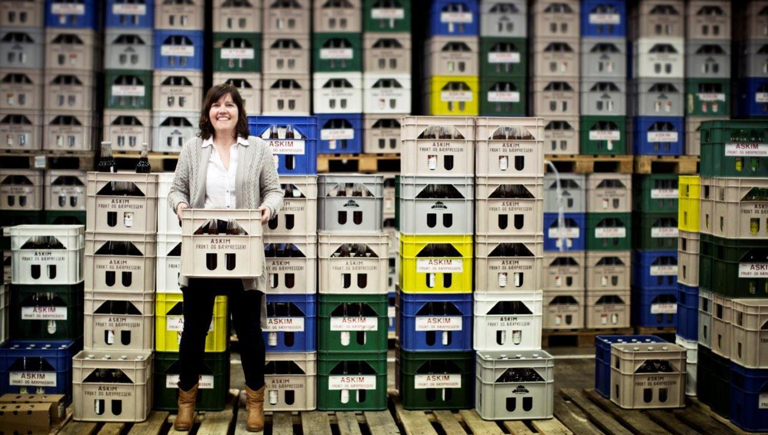 En kvinne står i et lager fullt av kasser stablet i høye rekker. Hun smiler og holder en kasse med flasker foran seg. Kassene har forskjellige farger og merkelapper, og rommet ser ryddig og organisert ut.