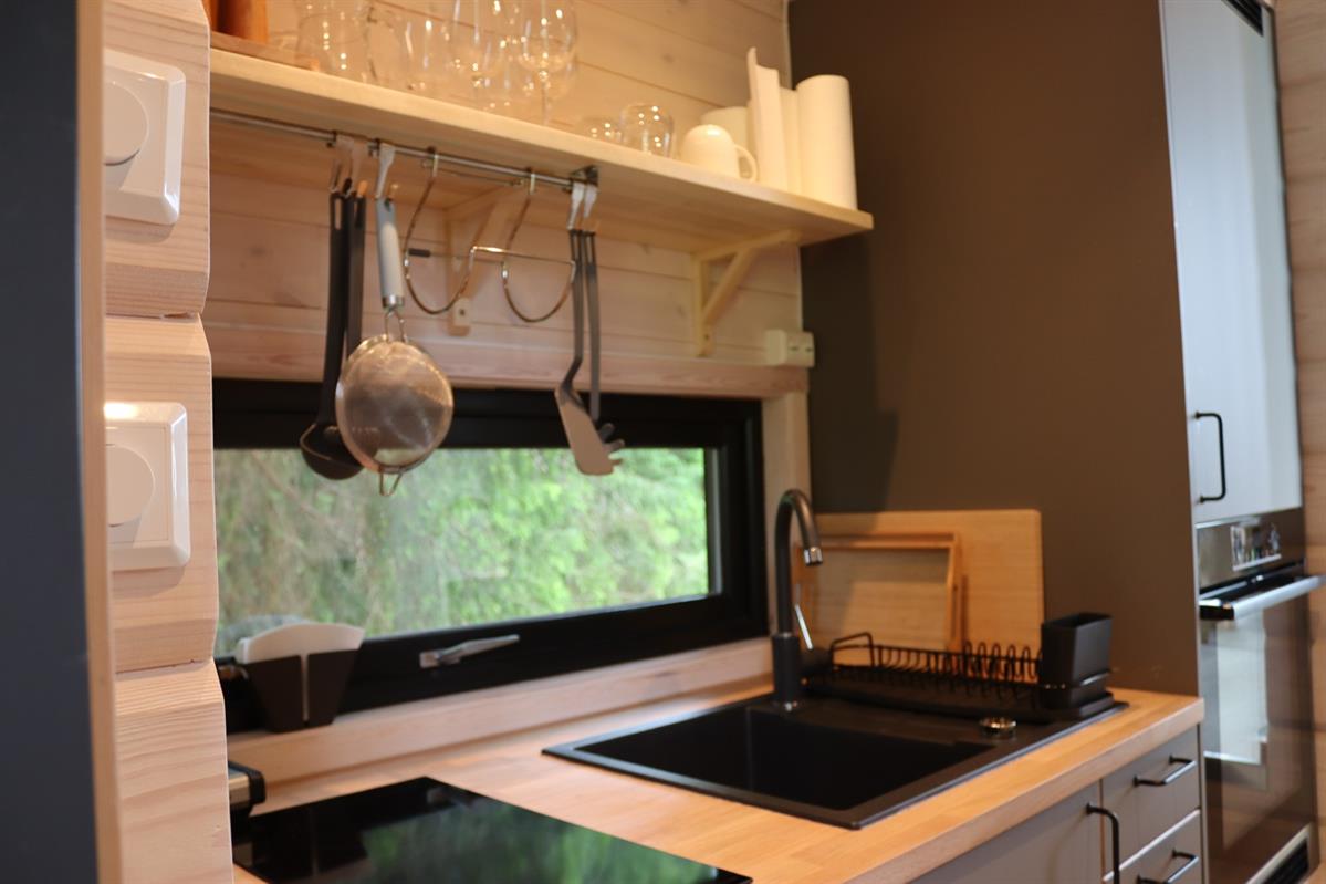 Small kitchen area with countertop, sink, and shelves holding kitchen utensils. A window offers a view of trees.
