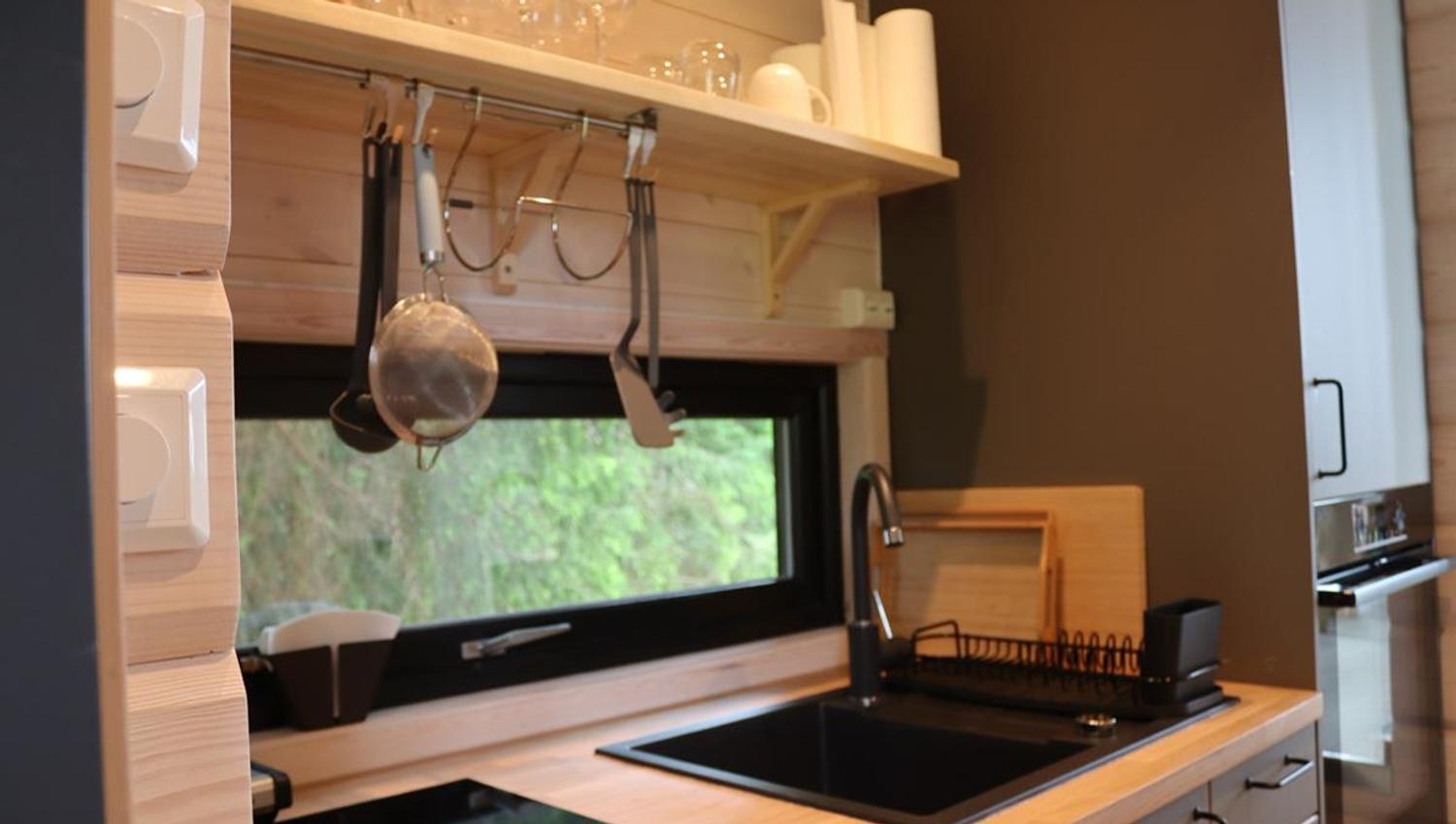 Small kitchen area with countertop, sink, and shelves holding kitchen utensils. A window offers a view of trees.