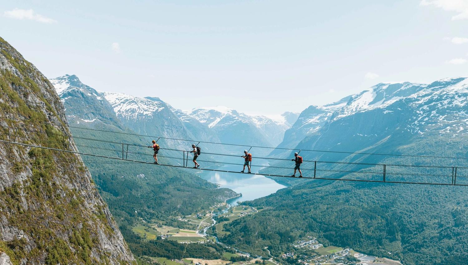 Fire personar med ryggsekk går på ei smal hengebru høgt over ein grøn dal, Lodalen, med fjord og snødekte fjell.