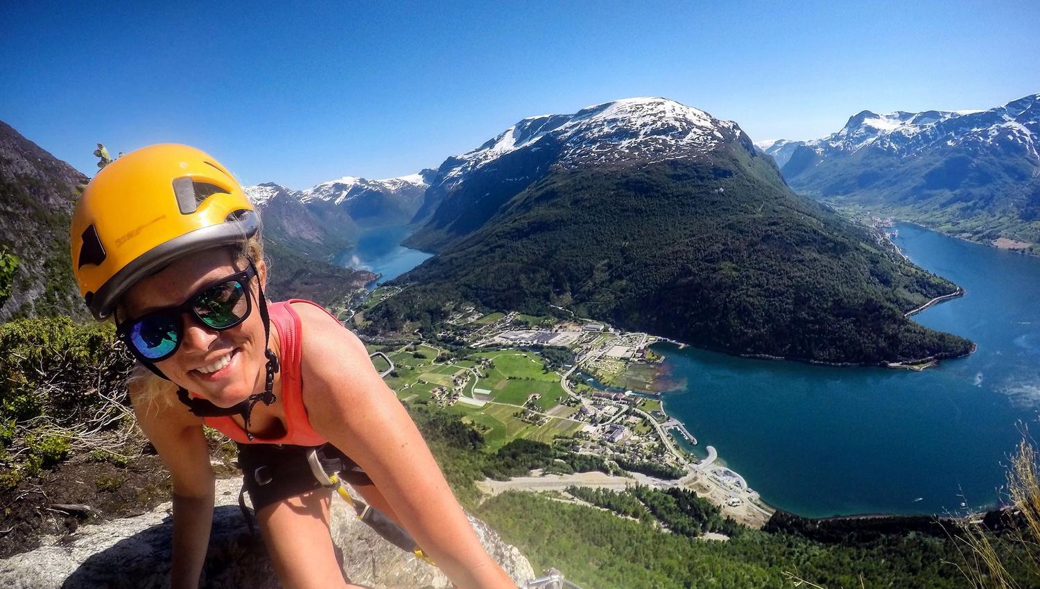 Ein klatrar med hjelm og sele på ein bratt fjellvegg, med fjord, bygder og snøkledde fjell i bakgrunnen..