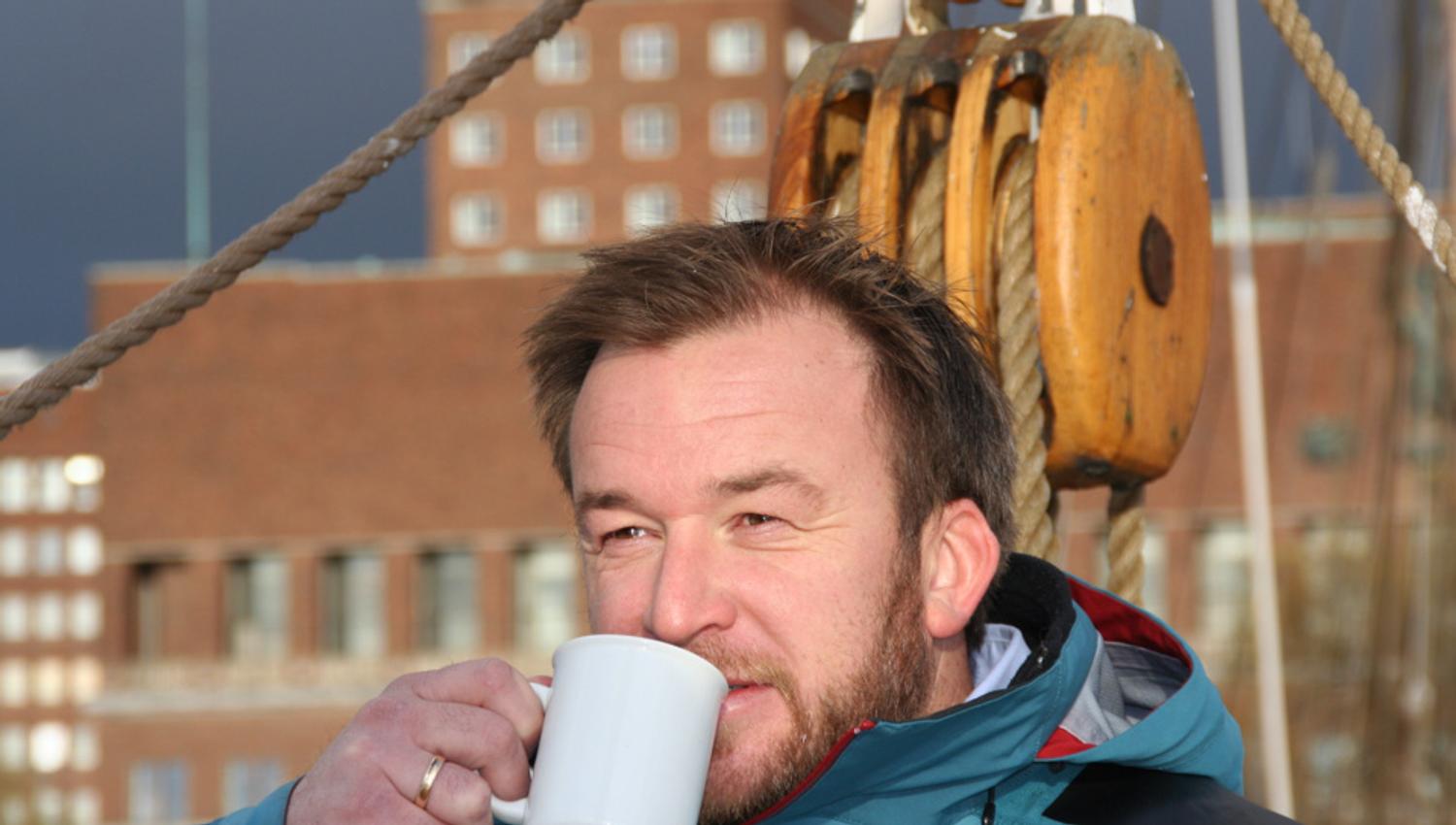 Man drinking coffee on deck with City Hall in the background