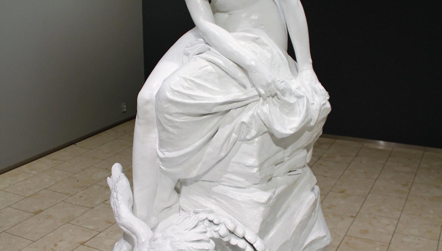 Hvit skulptur av en sittende naken kvinne på stein med en svane ved foten, utstilt i et gallerirom..