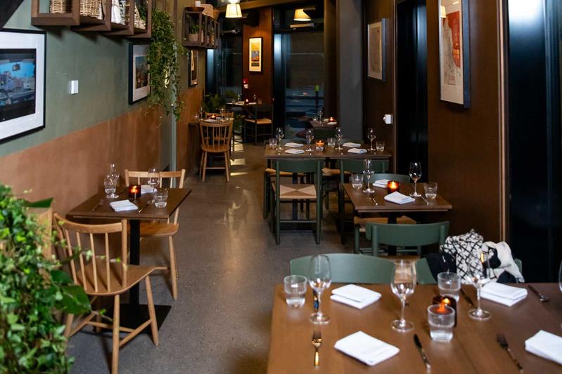 Restaurantinteriør med stor takhøyde, små dekkede trebord og stoler