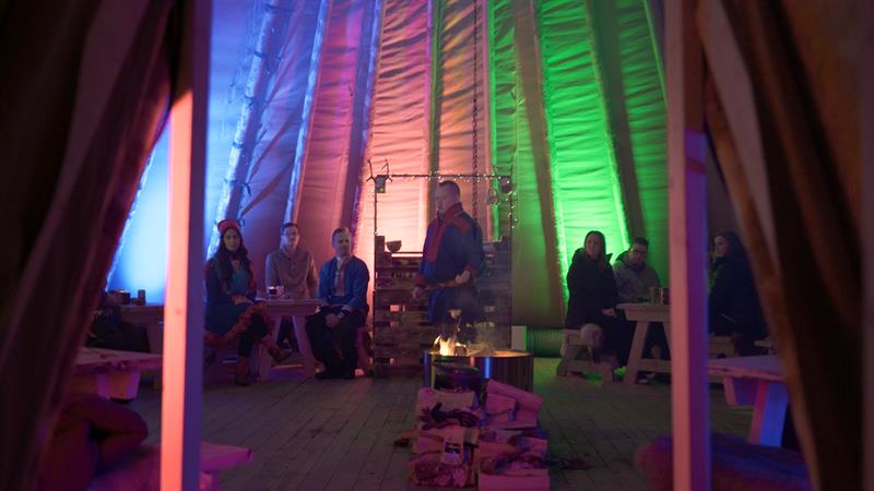 inside a lavvo with a bonfire and a man in front with people sitting around the fire 