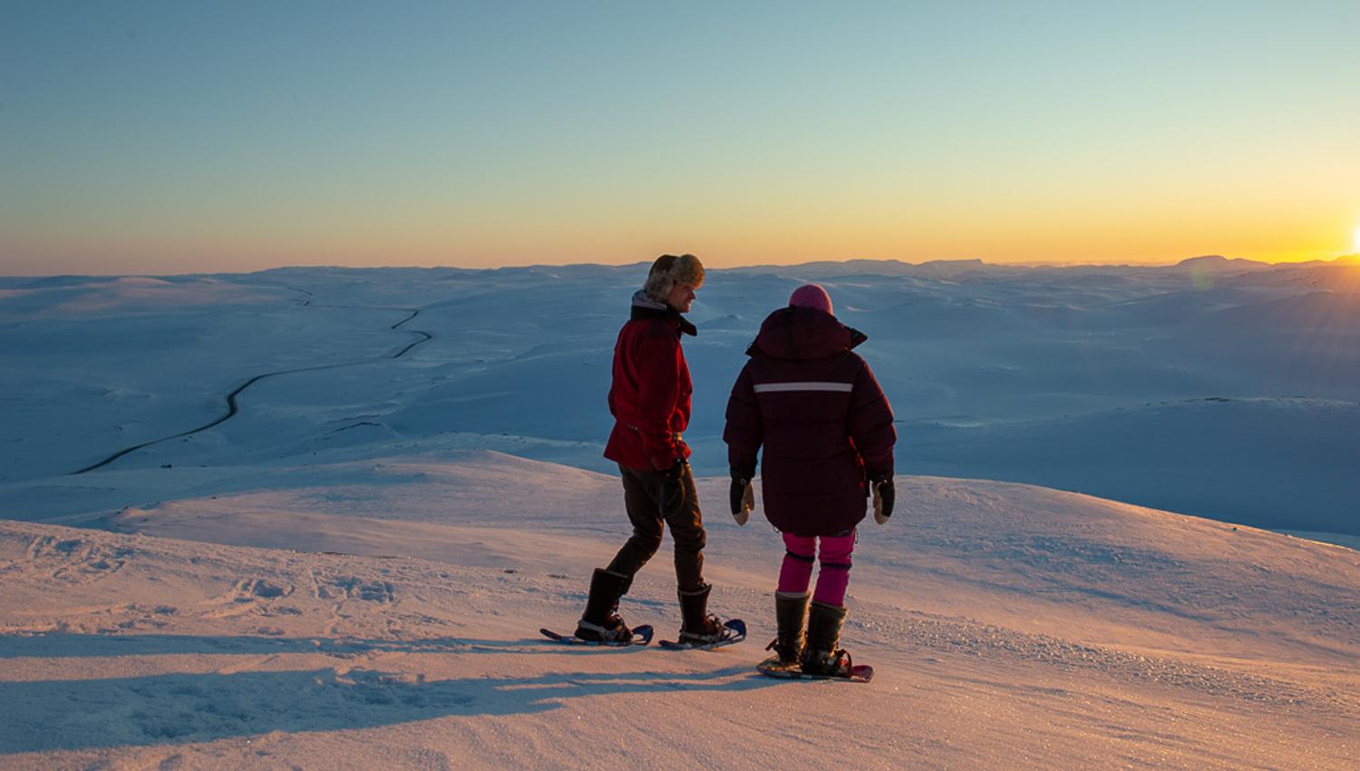 Two people on snowshoes on top of Halnekollen in a winter landscape at sunset.
