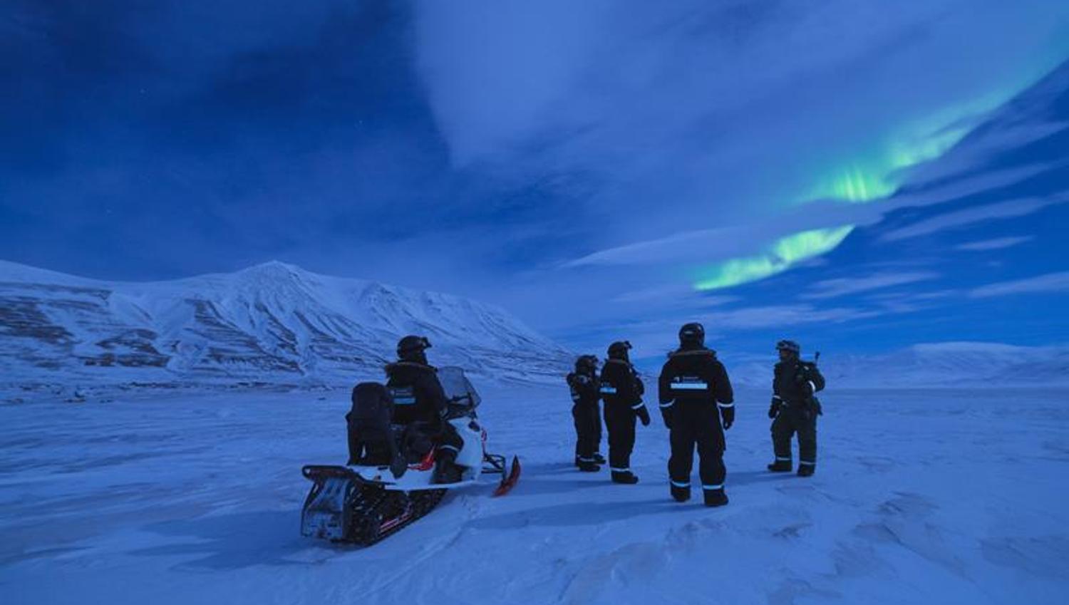 En gruppe mennesker og en snøscooter står ute i polarnatten. På himmelen er det nordlys.