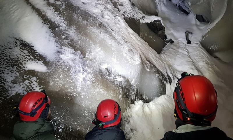 Tre personer med hodelykt og hjelm som beundrer formasjonene i taket på isgrotten.