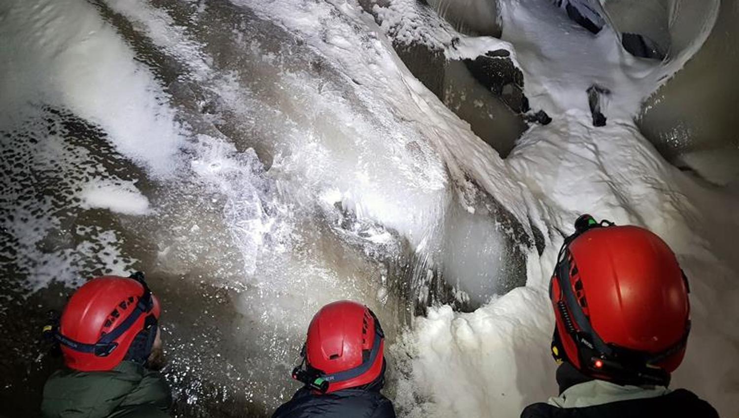 Tre personer med hodelykt og hjelm som beundrer formasjonene i taket på isgrotten.