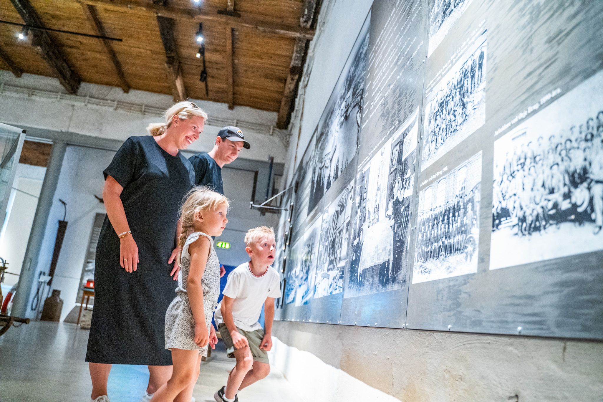 Bildet viser en familie, en kvinne, en mann og to barn som ser på store svart-hvite bilder på en museumsvegg. Alle smiler og virker interesserte. Rommet har høyt tak og store vinduer som slipper inn lys. Dette er på kanalmuseet.