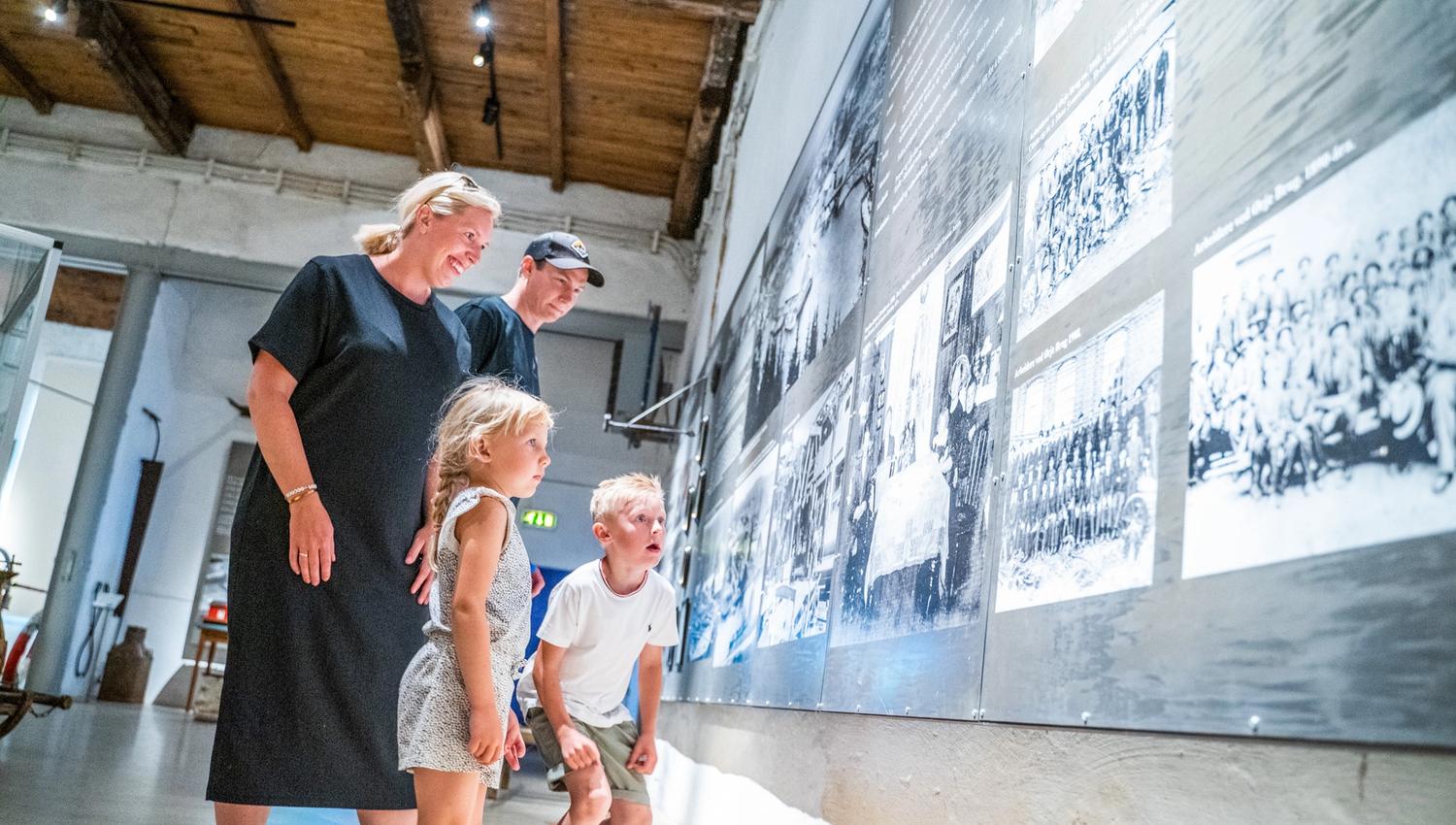 Bildet viser en familie, en kvinne, en mann og to barn som ser på store svart-hvite bilder på en museumsvegg. Alle smiler og virker interesserte. Rommet har høyt tak og store vinduer som slipper inn lys. Dette er på kanalmuseet.