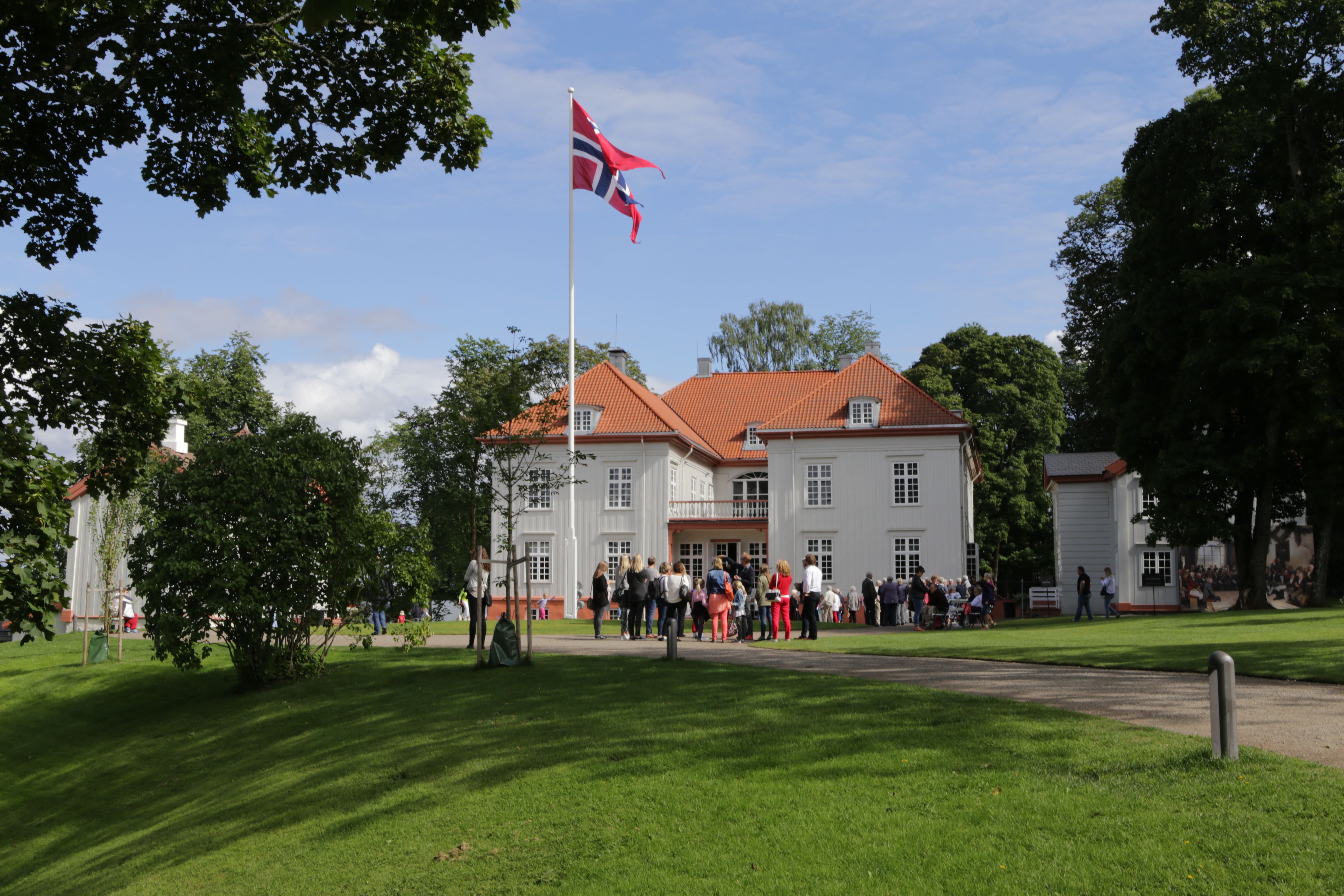 Eidsvoll bygningen i sommervær med vaiende flagg og mange mennesker utenfor, grønnkledde trær og gress.