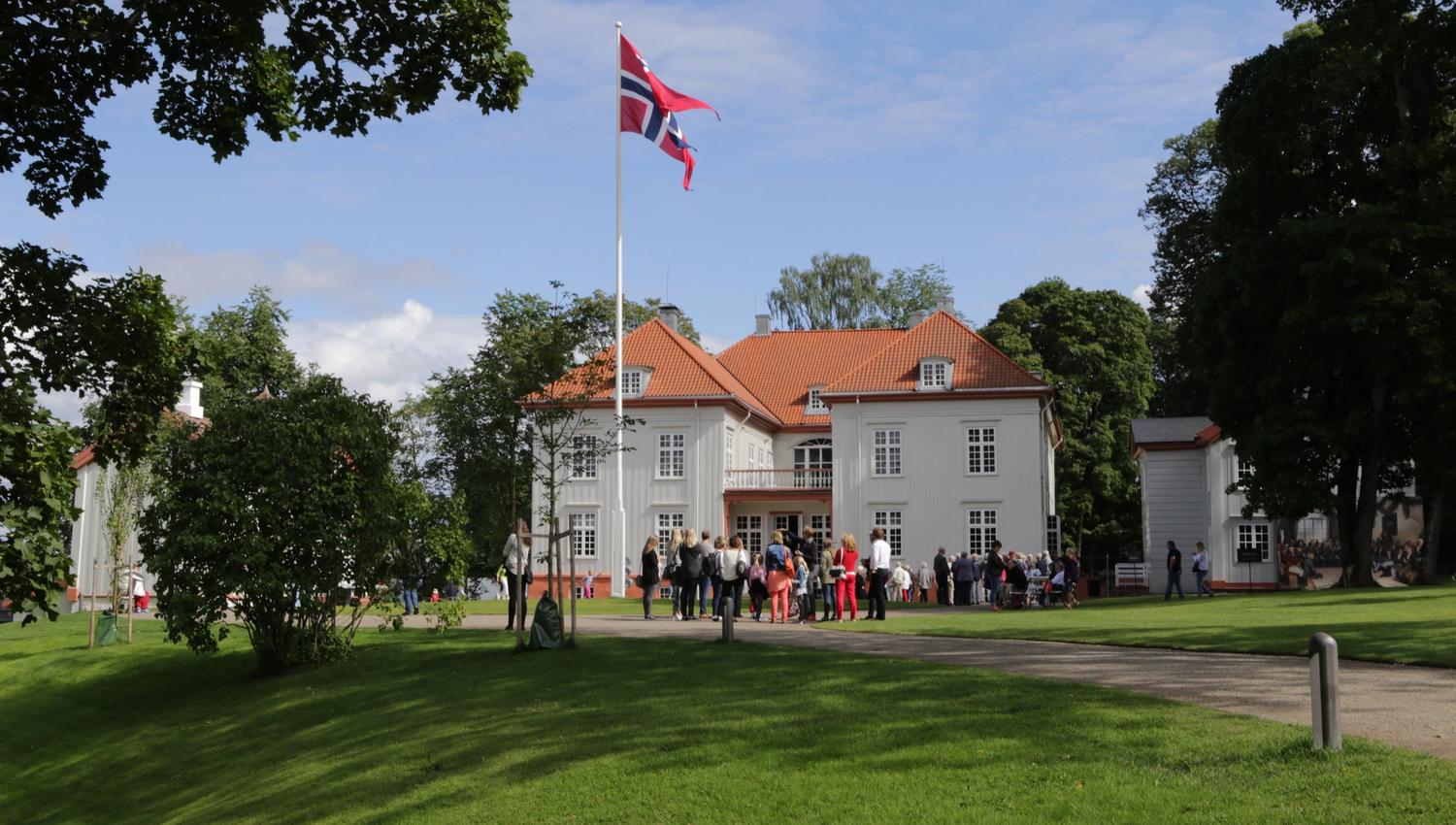 Eidsvoll bygningen i sommervær med vaiende flagg og mange mennesker utenfor, grønnkledde trær og gress.