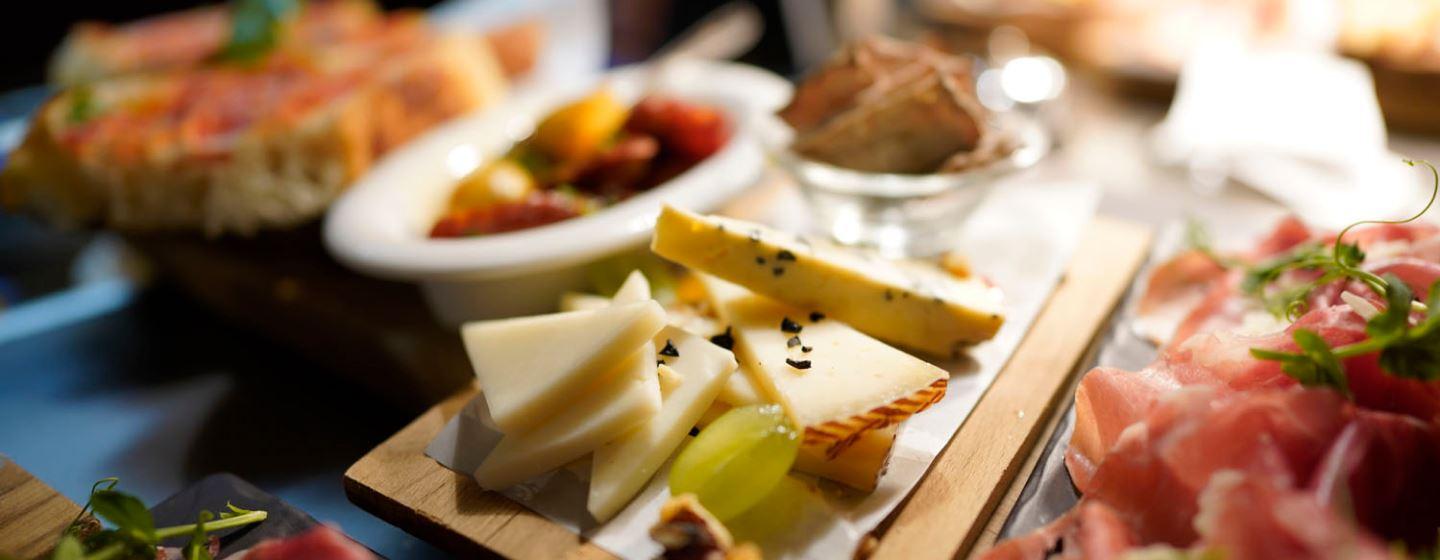 Close-up of cheese with black seeds, green grapes and crackers, with cured meats and bruschetta in the background.