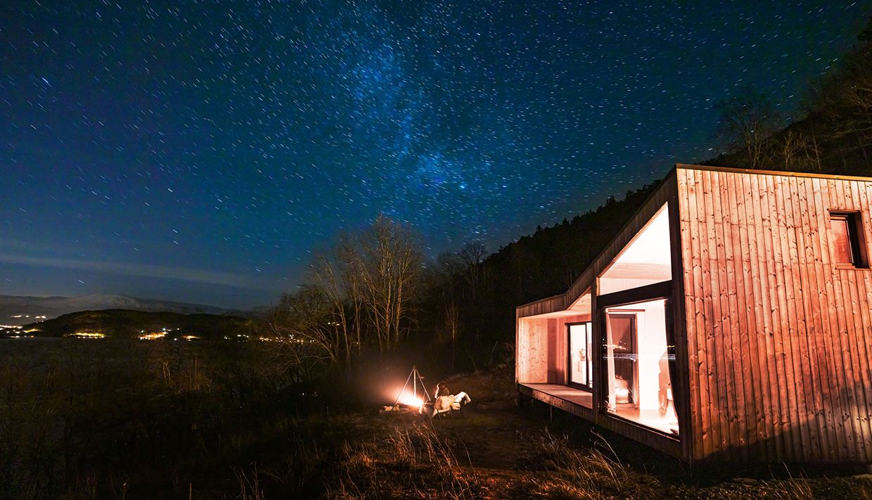 Modern cabin under a starry sky by Hardangerfjord, with a campfire outside.