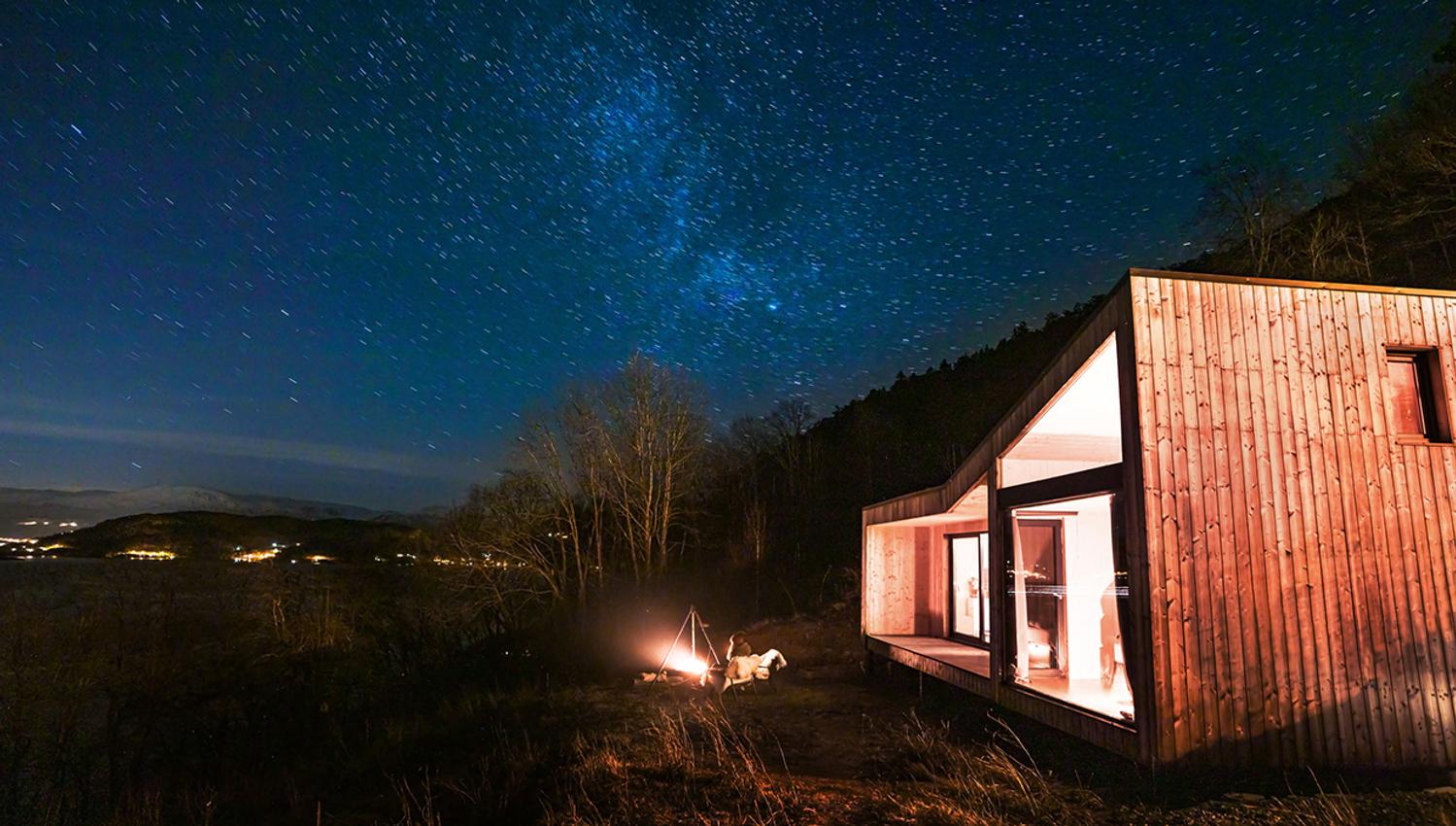 Modern cabin under a starry sky by Hardangerfjord, with a campfire outside.