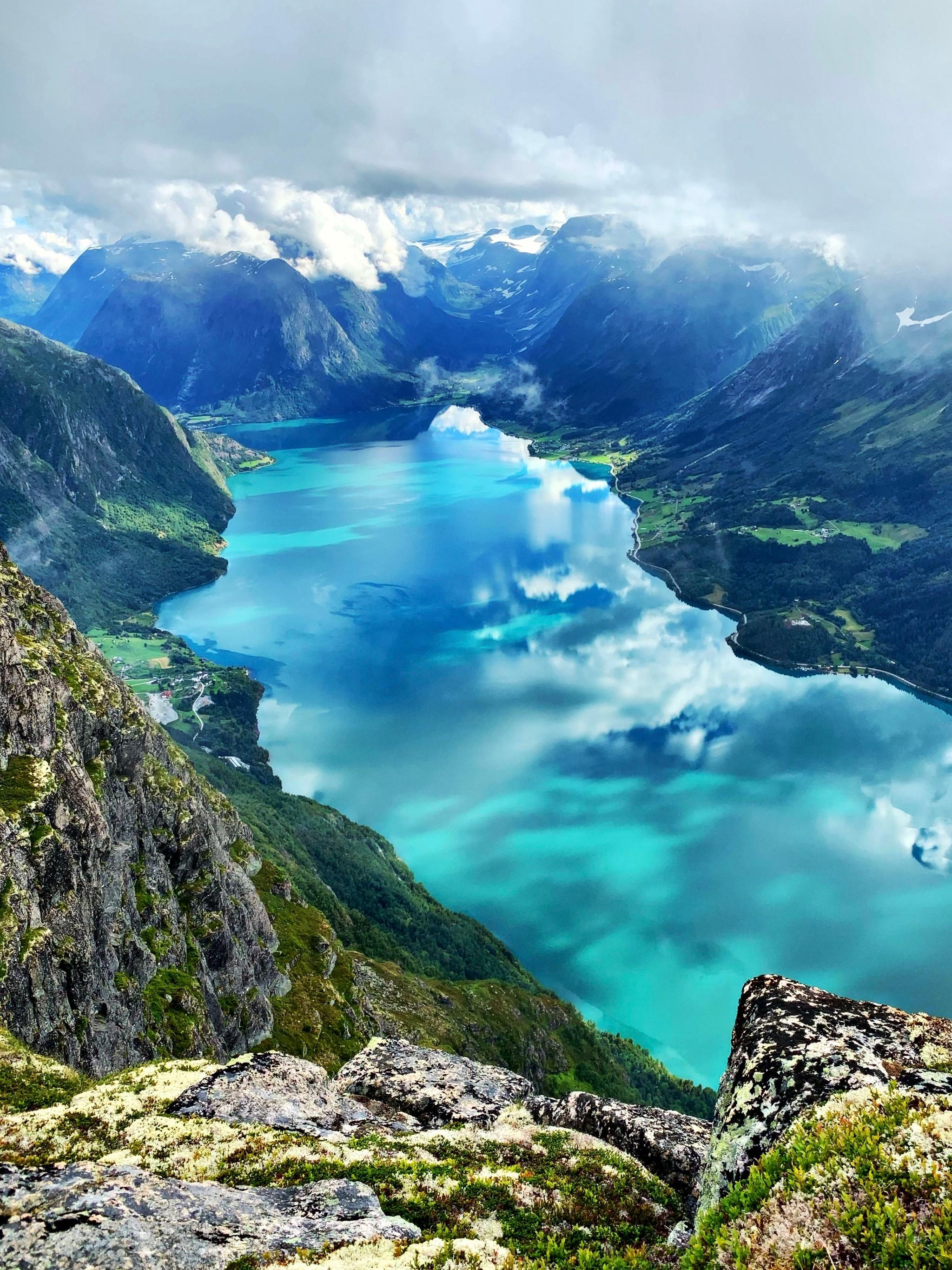 High viewpoint over a turquoise fjord surrounded by steep green mountains, rocky foreground, and low clouds..