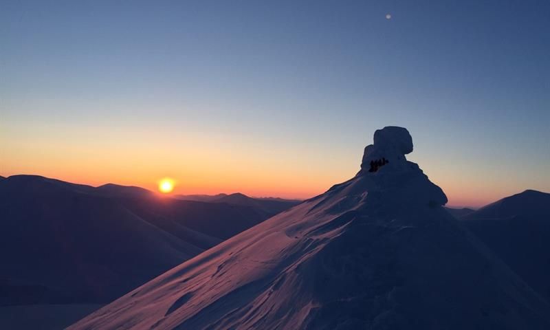 En snødekt fjelltopp i forgrunnen med en lav sol og klar himmel på horisonten over andre fjelltopper i bakgrunnen