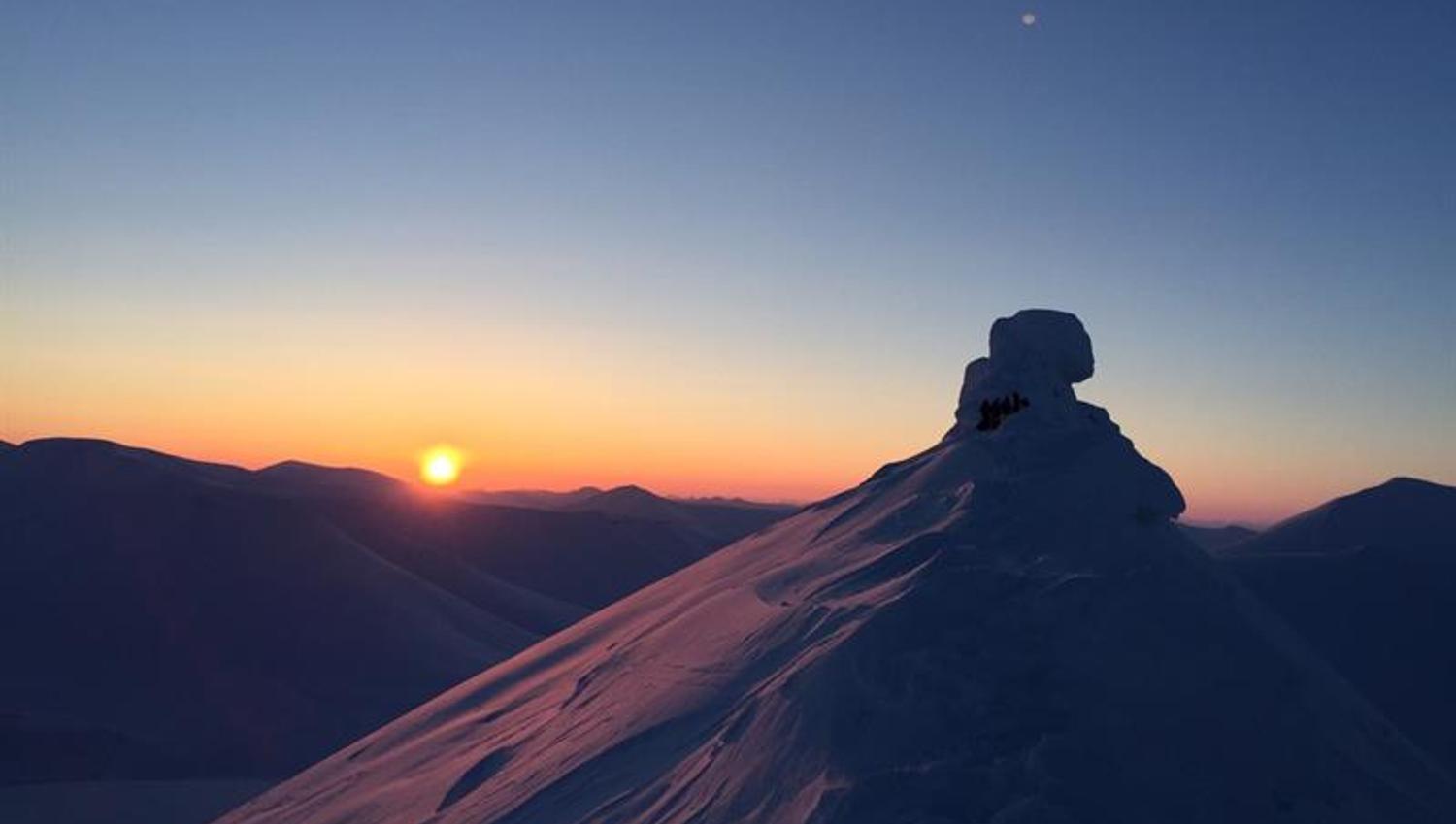 En snødekt fjelltopp i forgrunnen med en lav sol og klar himmel på horisonten over andre fjelltopper i bakgrunnen