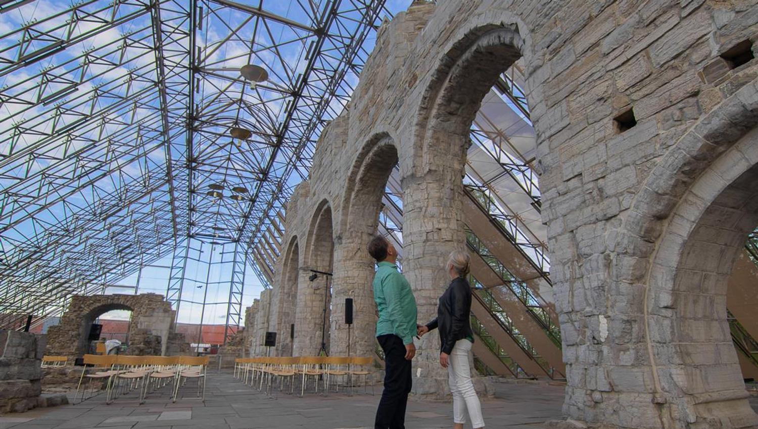 Interior view of the Hamardomen protective structure, showing tall glass walls and wooden beams surrounding the cathedral ruins. The natural light giv