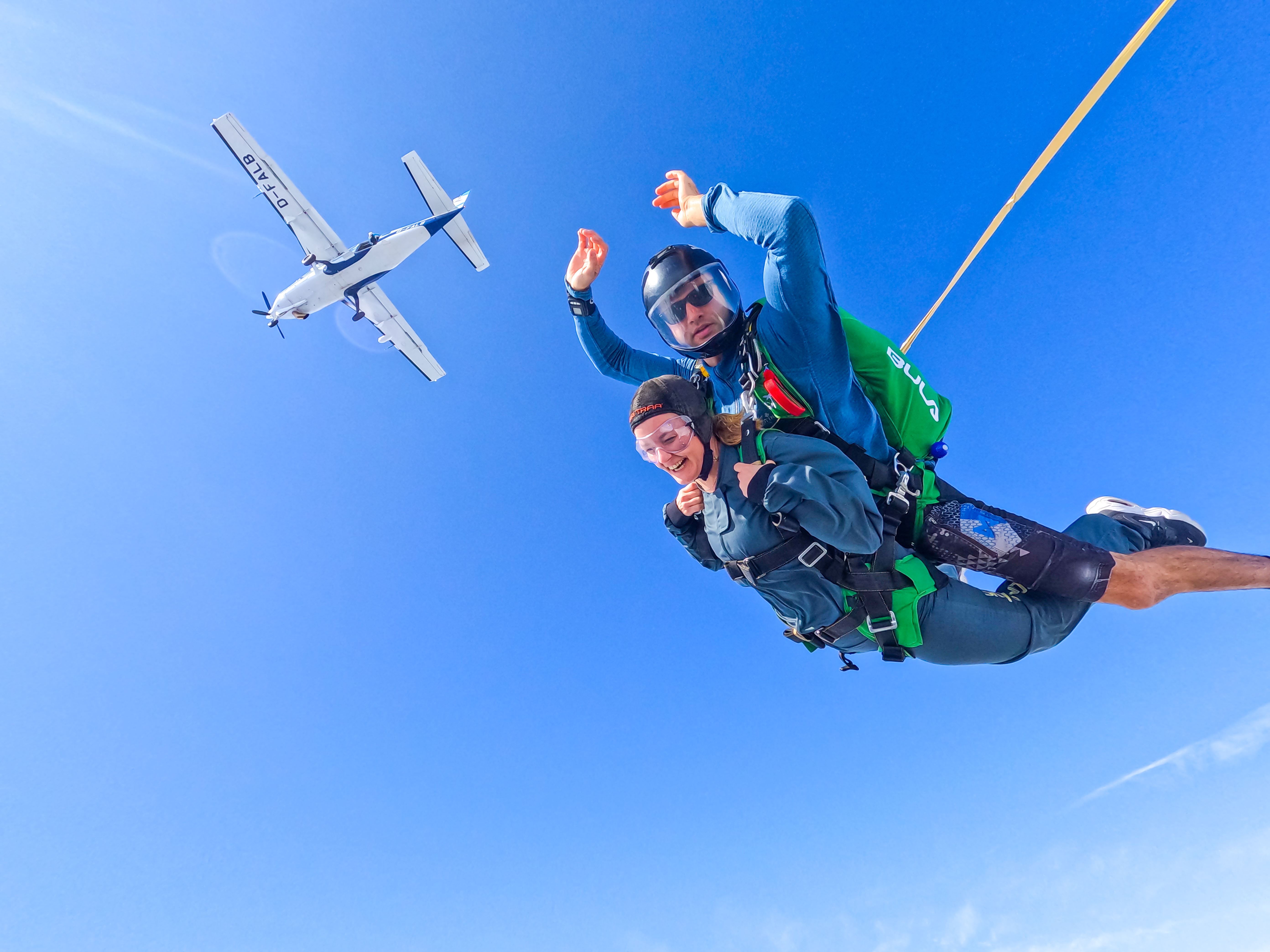 Tandem skydiving instructor and student in freefall, holding stable body position. Clear skies and the plane in the background, above them. 
