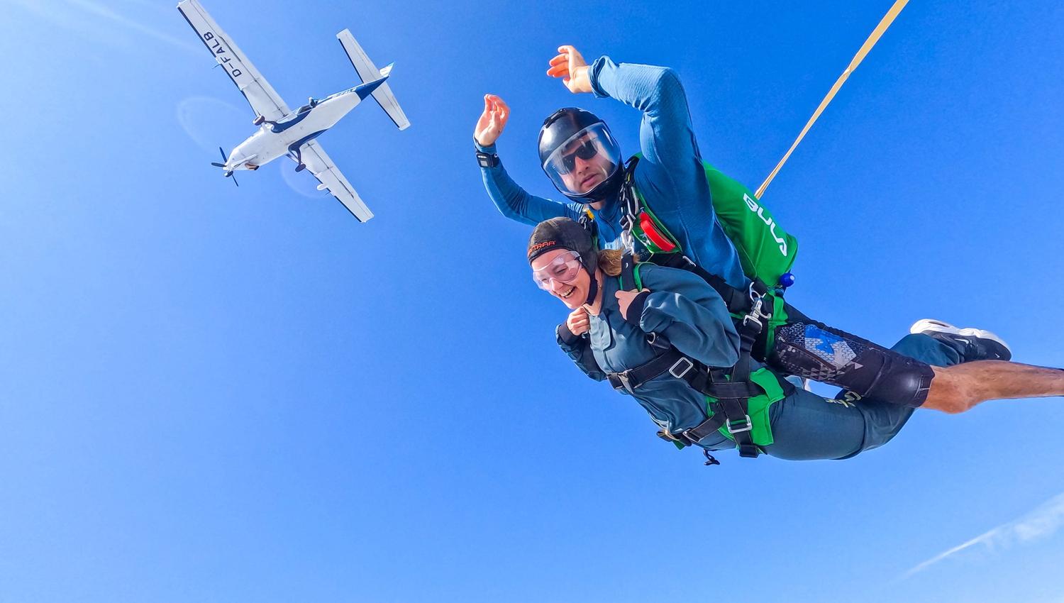 Tandem skydiving instructor and student in freefall, holding stable body position. Clear skies and the plane in the background, above them.