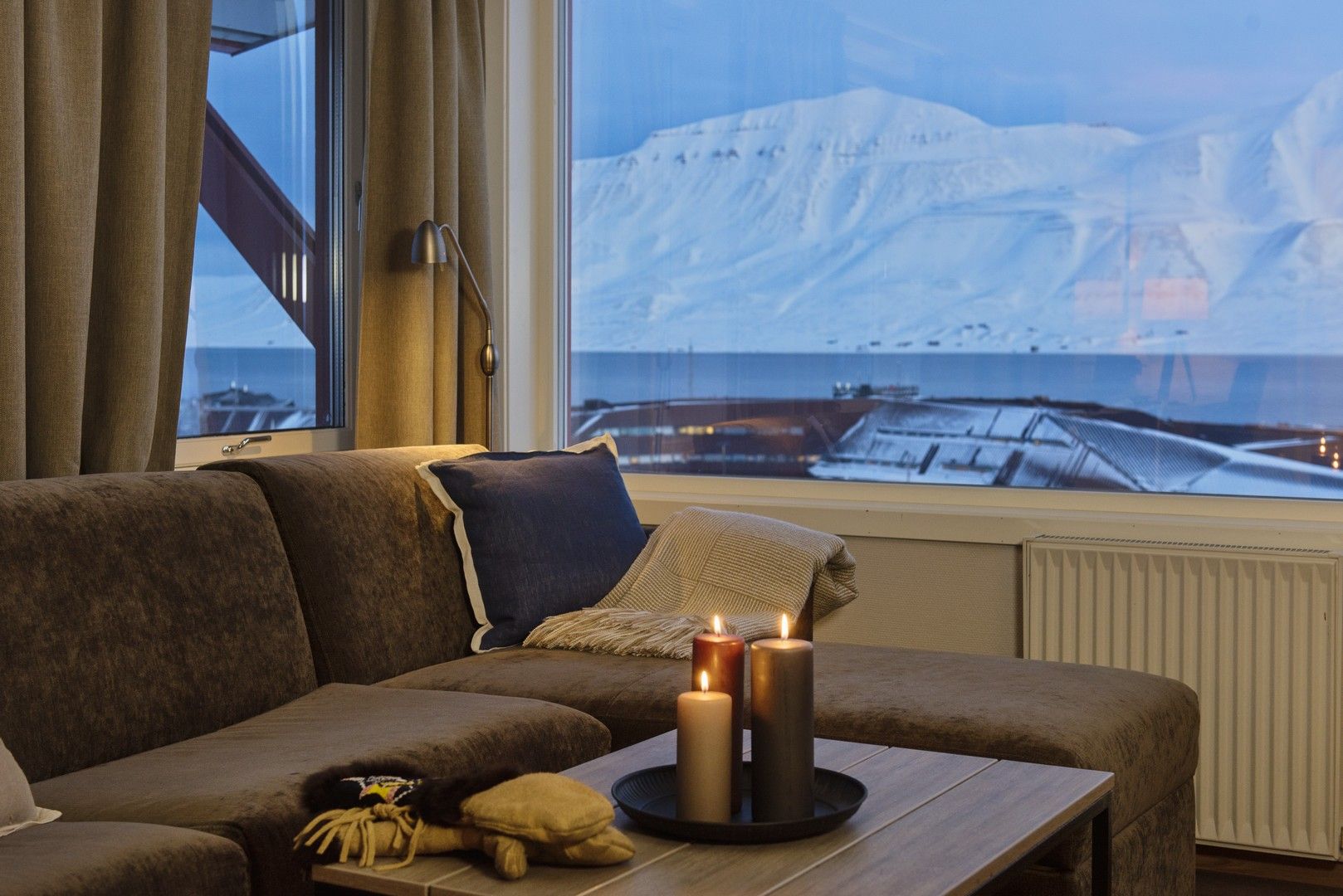 A couch with candlelights on a coffee table and a view towards snow-covered mountains in a blue light in the background