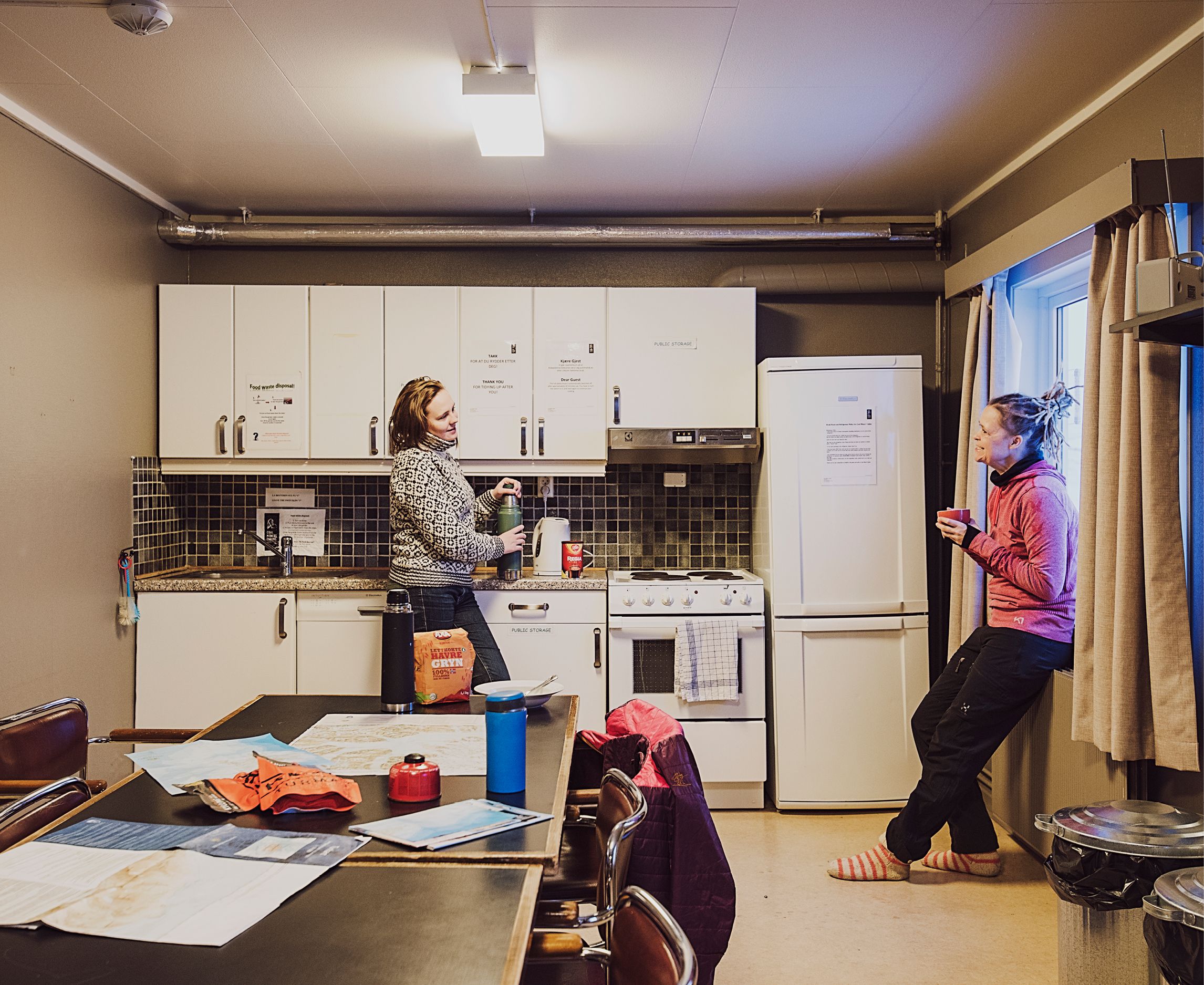 Two guests talking with each other in a shared kitchen area at Coal Miners' Cabins, with a table in the foreground on which various outdoors equipment