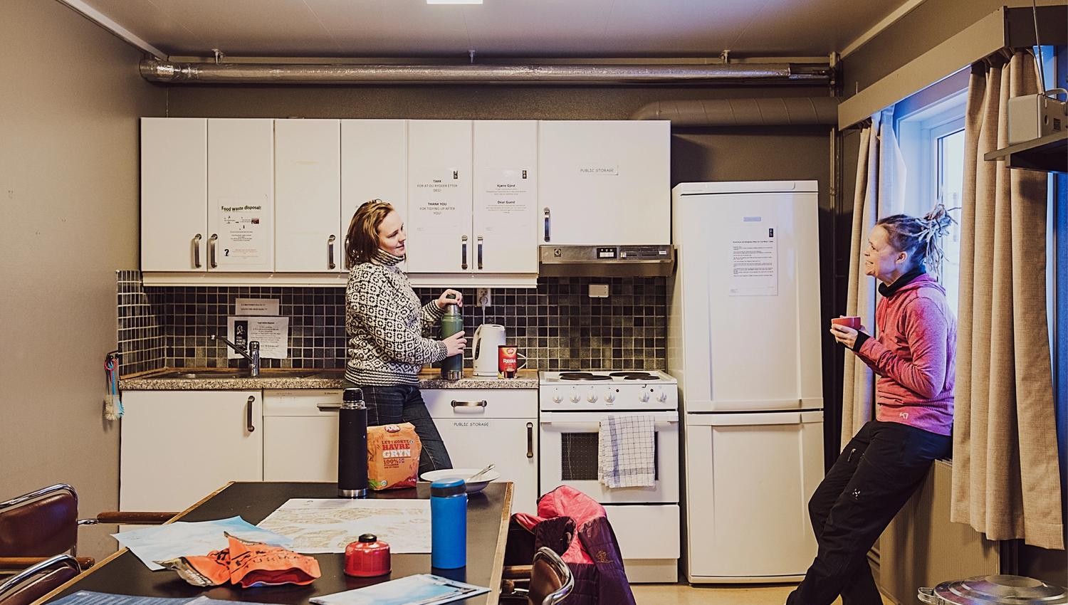 Two guests talking with each other in a shared kitchen area at Coal Miners' Cabins, with a table in the foreground on which various outdoors equipment
