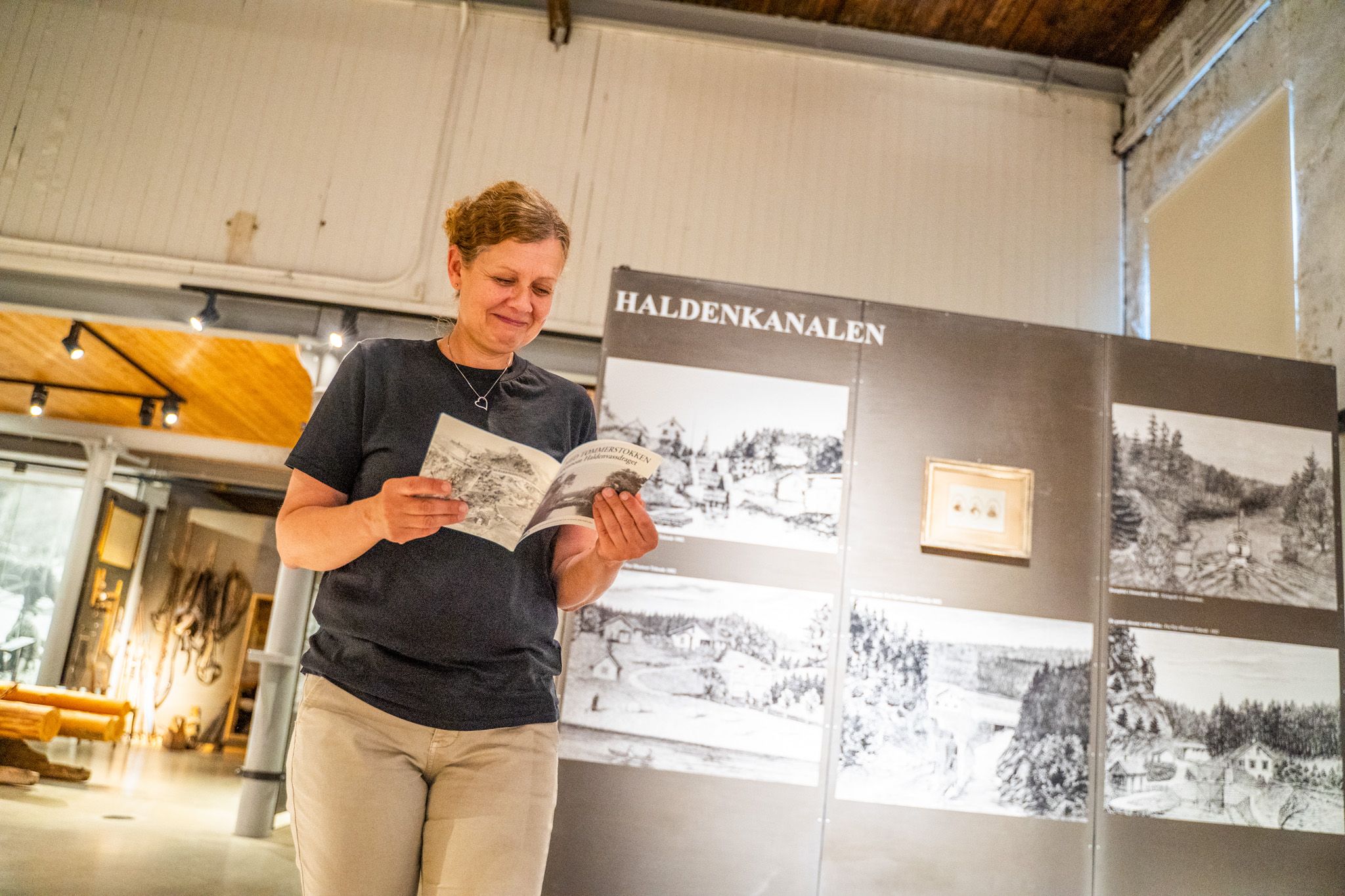 Bildet viser en kvinne som står og leser en brosjyre inne på et museum. Bak henne er en utstilling med bilder og tekst om Haldenkanalen. Hun smiler og virker interessert i det hun leser.