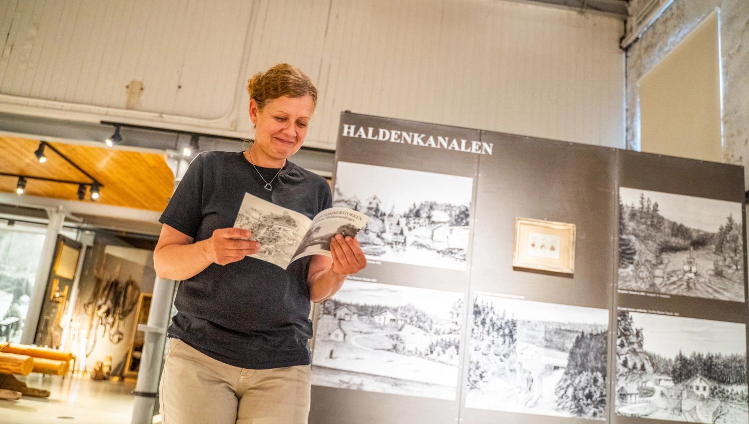 Bildet viser en kvinne som står og leser en brosjyre inne på et museum. Bak henne er en utstilling med bilder og tekst om Haldenkanalen. Hun smiler og virker interessert i det hun leser.