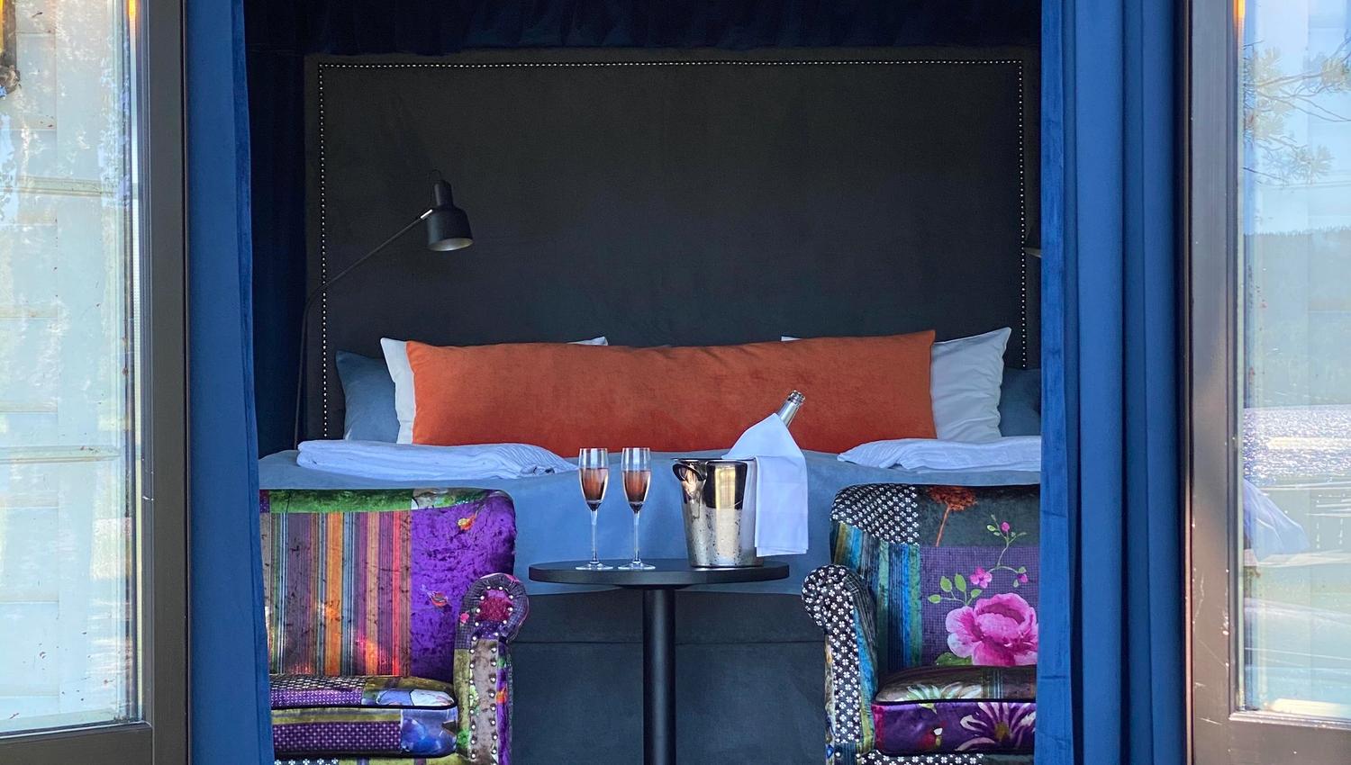 2 stoler med blomstrete trekk, 2 champagne glass på et bord foran en seng i et rom sett utenfra