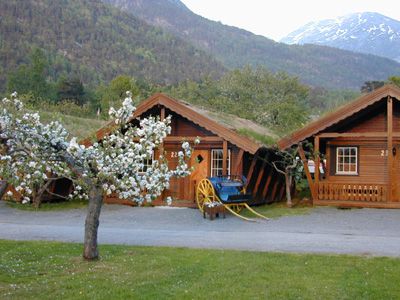 Sjarmerande tømmerhytter i vakre omgjevnader på Mikkelparken Ferietun, med blomstrande frukttre og fjell i bakgrunnen.