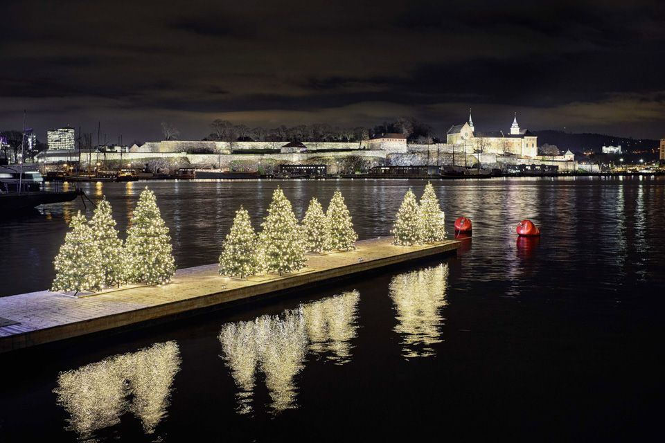 Nattbilde av brygge med juletær med tente lys på. Akershus festning i bakgrunnen.