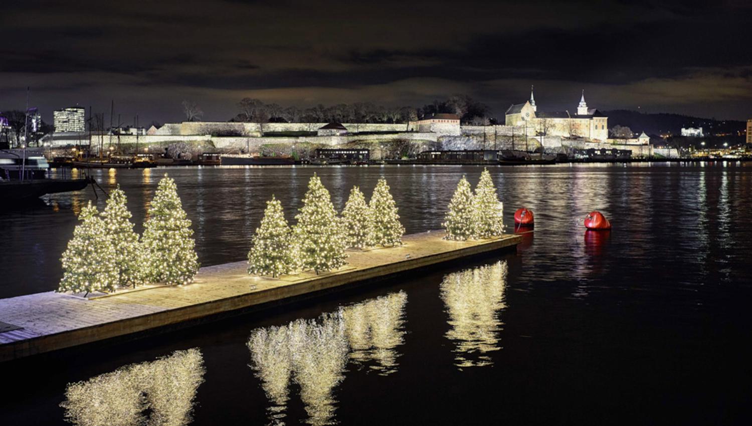 Nattbilde av brygge med juletær med tente lys på. Akershus festning i bakgrunnen.