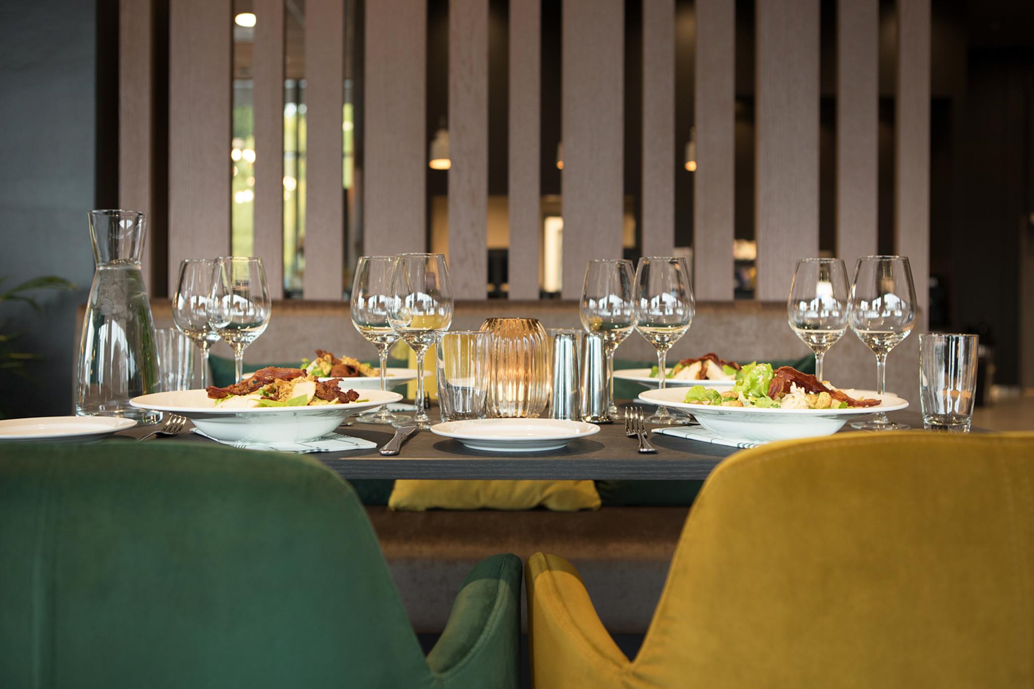 Bildet viser et pent dekket bord på en restaurant eller et moderne spisested. Det står tallerkener med mat, mange vinglass, vannglass og bestikk på bordet. Stolene er i forskjellige farger – en grønn og en gul – og stemningen virker rolig og elegant.