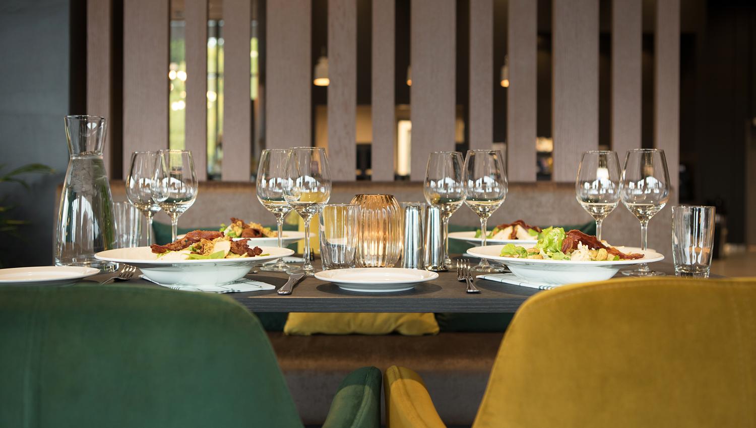 Bildet viser et pent dekket bord på en restaurant eller et moderne spisested. Det står tallerkener med mat, mange vinglass, vannglass og bestikk på bordet. Stolene er i forskjellige farger – en grønn og en gul – og stemningen virker rolig og elegant.