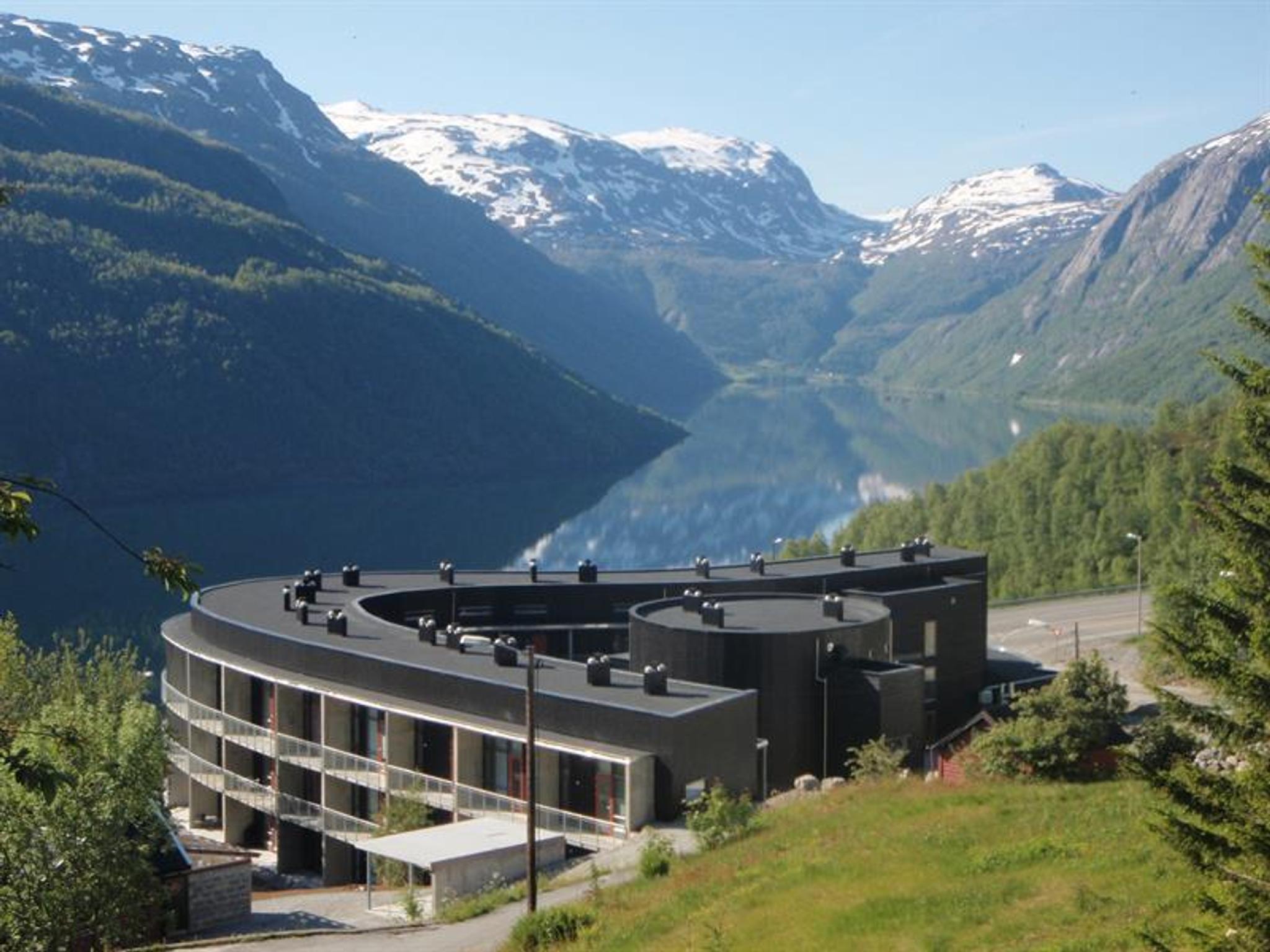 Hordatun Hotel sett frå sommarlandskap, med utsikt mot Røldalsvatnet og dei snødekte fjelltoppane i bakgrunnen.