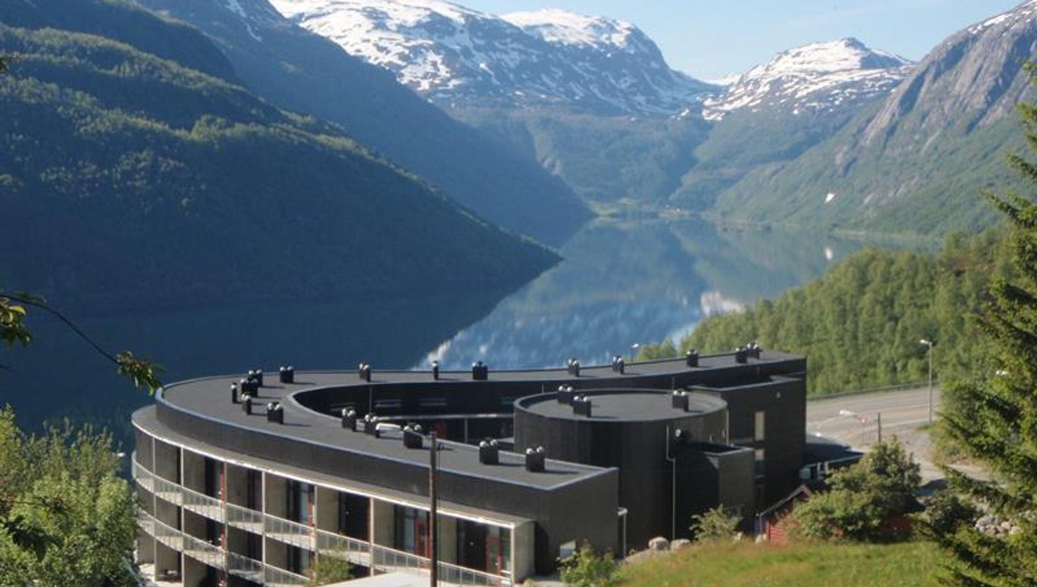 Hordatun Hotel sett frå sommarlandskap, med utsikt mot Røldalsvatnet og dei snødekte fjelltoppane i bakgrunnen.