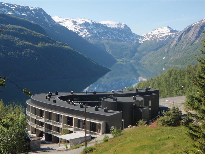 Hordatun Hotel sett frå sommarlandskap, med utsikt mot Røldalsvatnet og dei snødekte fjelltoppane i bakgrunnen.