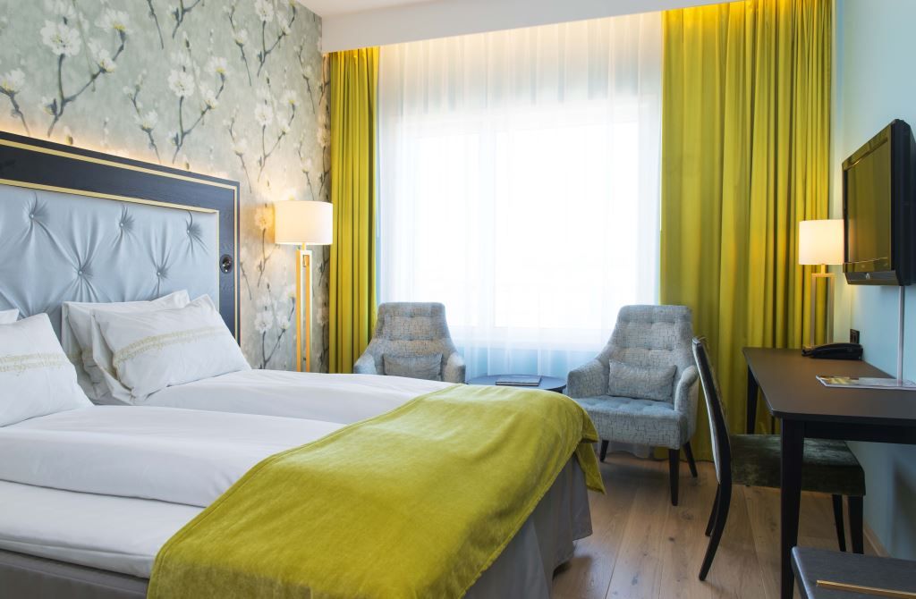 Interior of hotel room: Double bed, blue armchairs, TV screen on the wall, floral wallpaper.