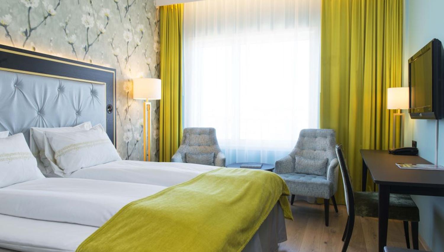 Interior of hotel room: Double bed, blue armchairs, TV screen on the wall, floral wallpaper.