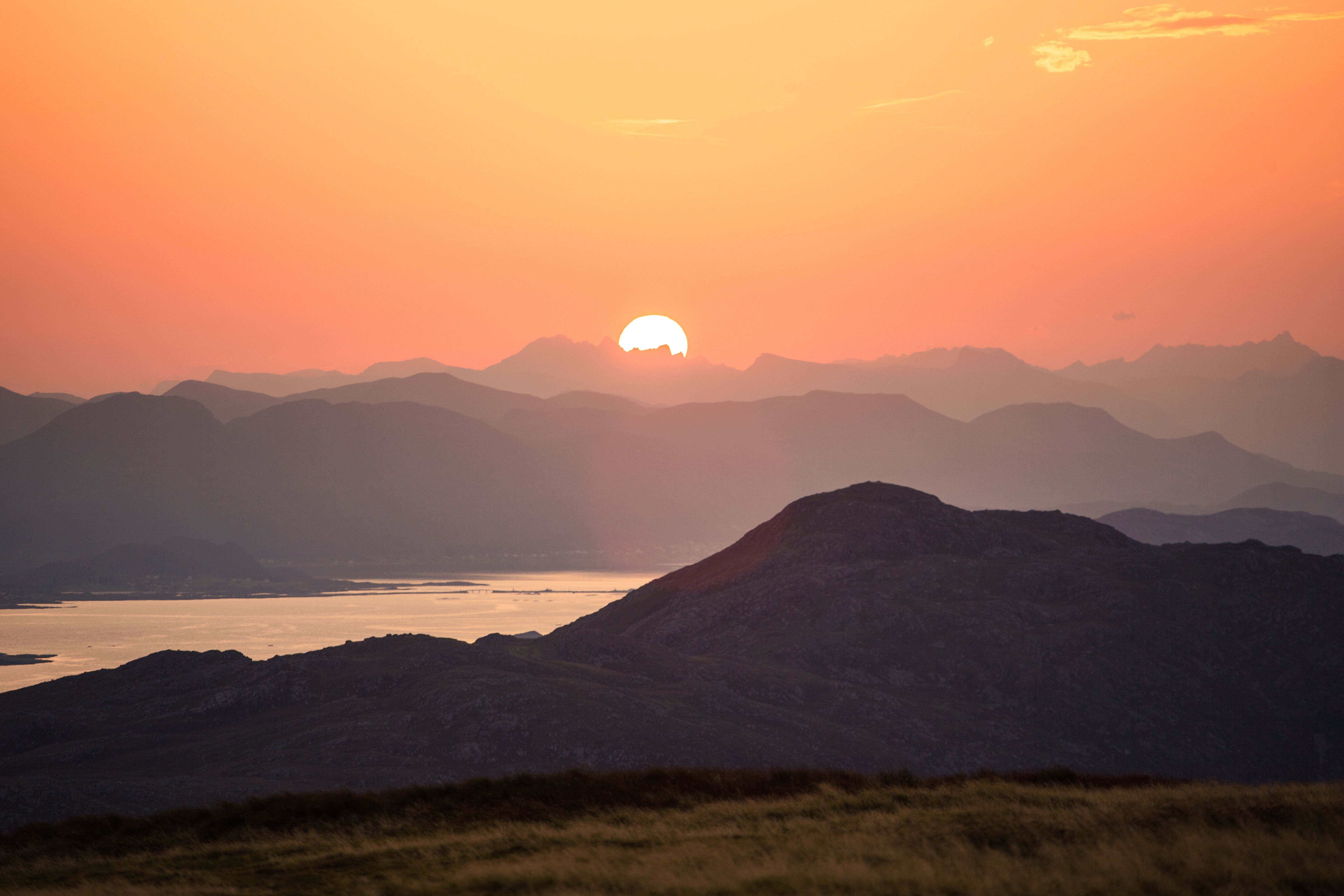 Sol går ned bak lag på lag med fjell, med varm oransje himmel og spegling i stille fjordvatn..