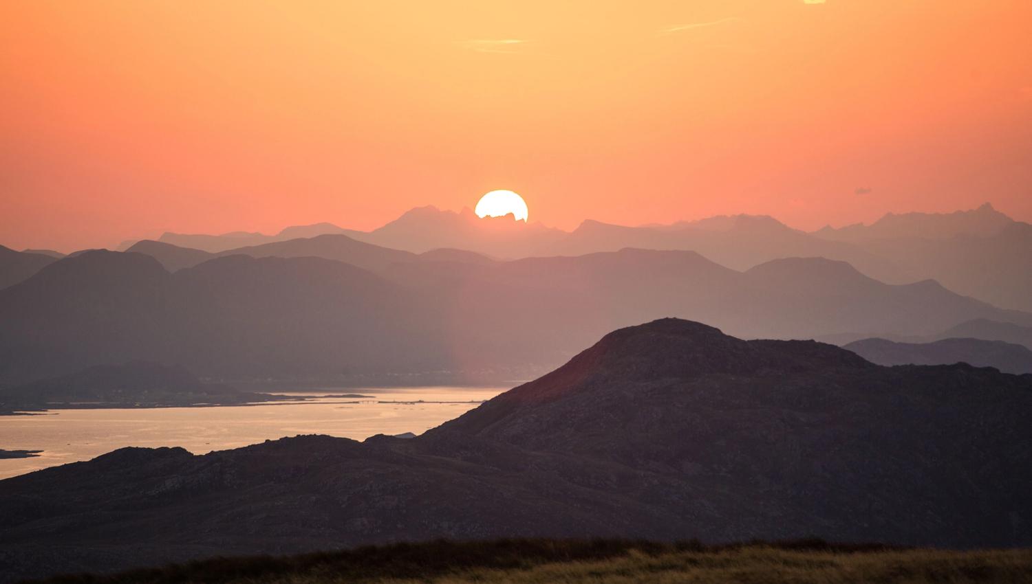 Sol går ned bak lag på lag med fjell, med varm oransje himmel og spegling i stille fjordvatn..