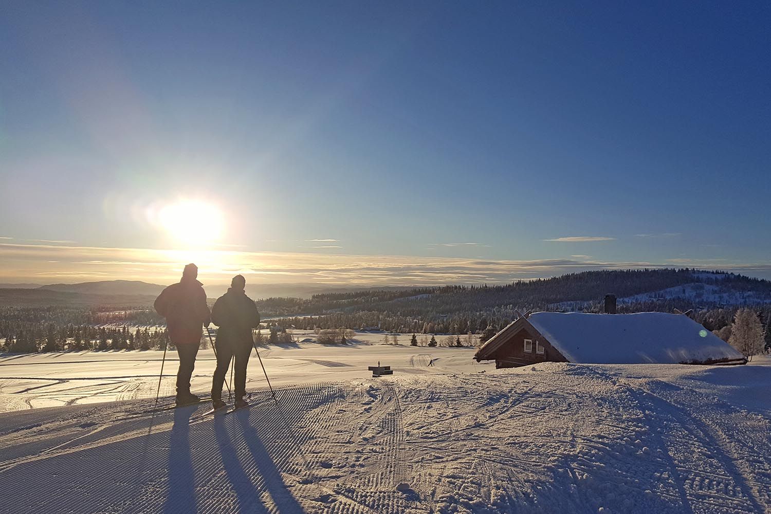 Two people in a cross-country skiing track by a cabin against the setting sun