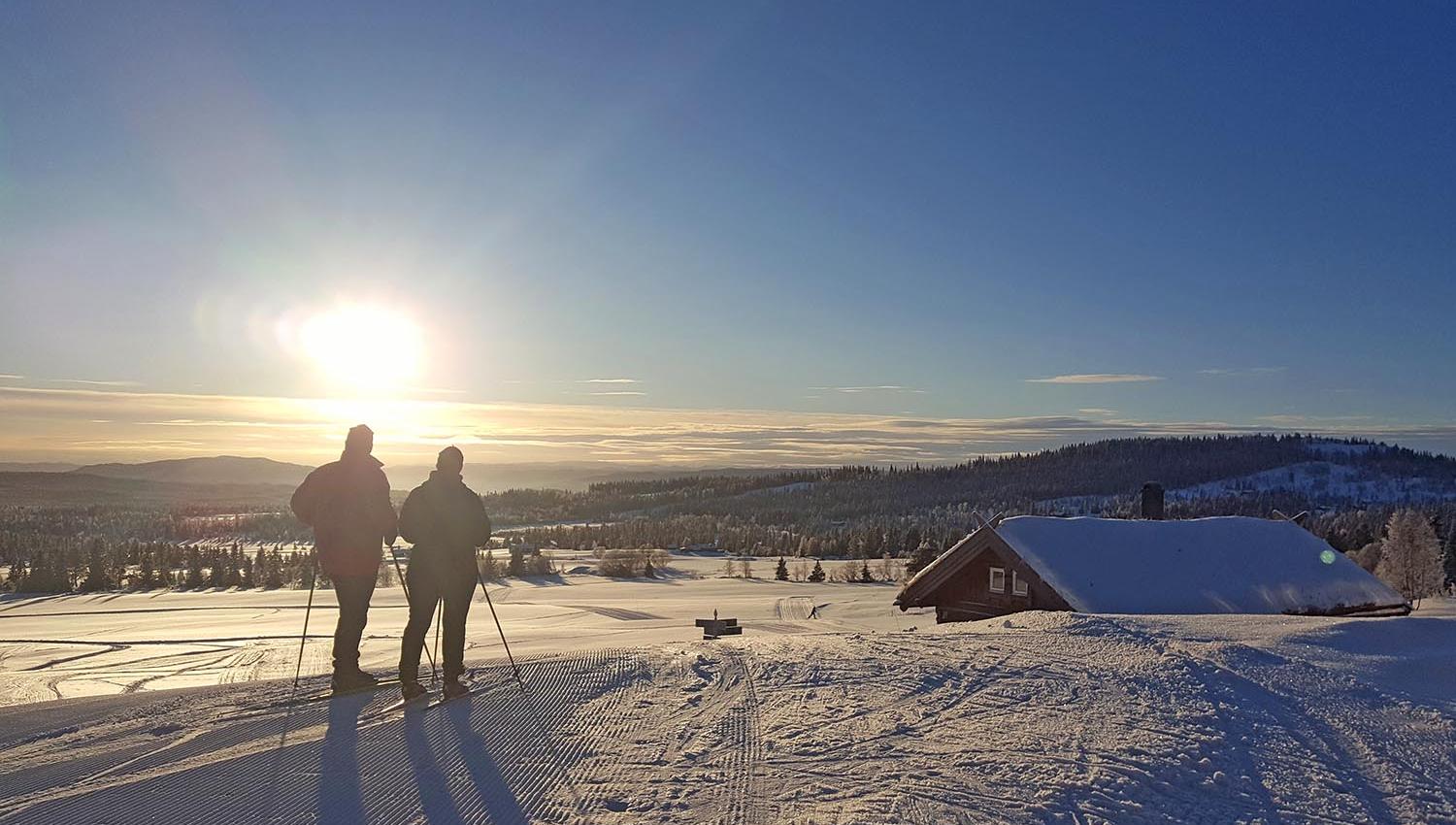 Two people in a cross-country skiing track by a cabin against the setting sun