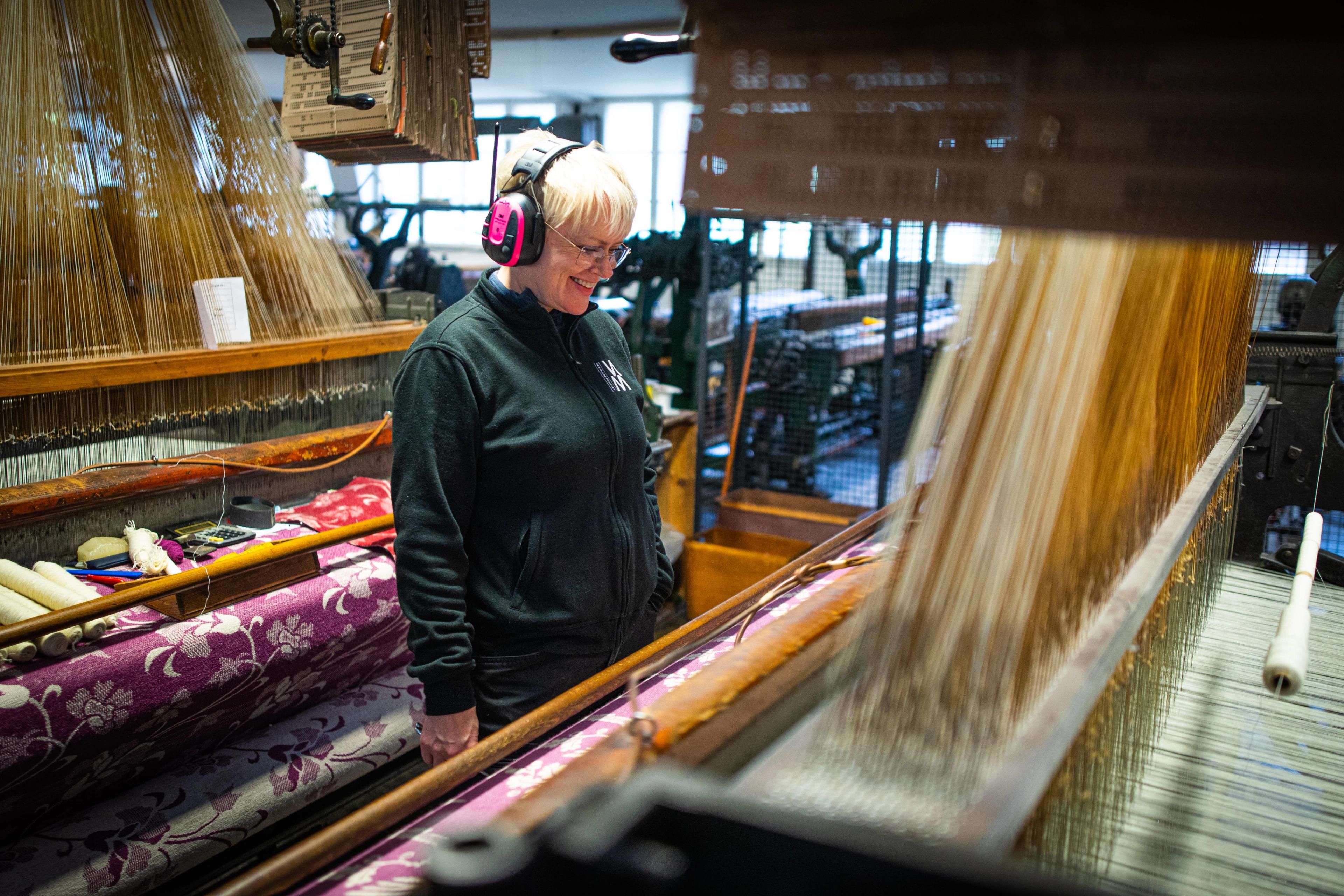 Smiling woman wearing pink hearing protection, standing and watching the weaving machine.