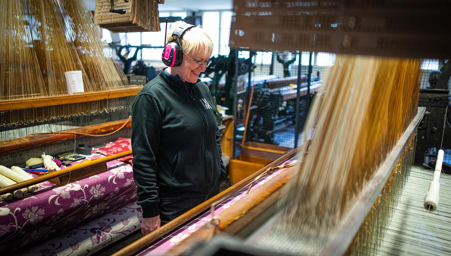 Smiling woman wearing pink hearing protection, standing and watching the weaving machine.