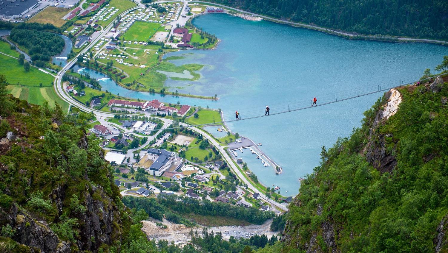 Utsyn frå fjellside mot ein fjord og ei bygd, med ei smal hengebru der fleire personar kryssar høgt over vatnet..