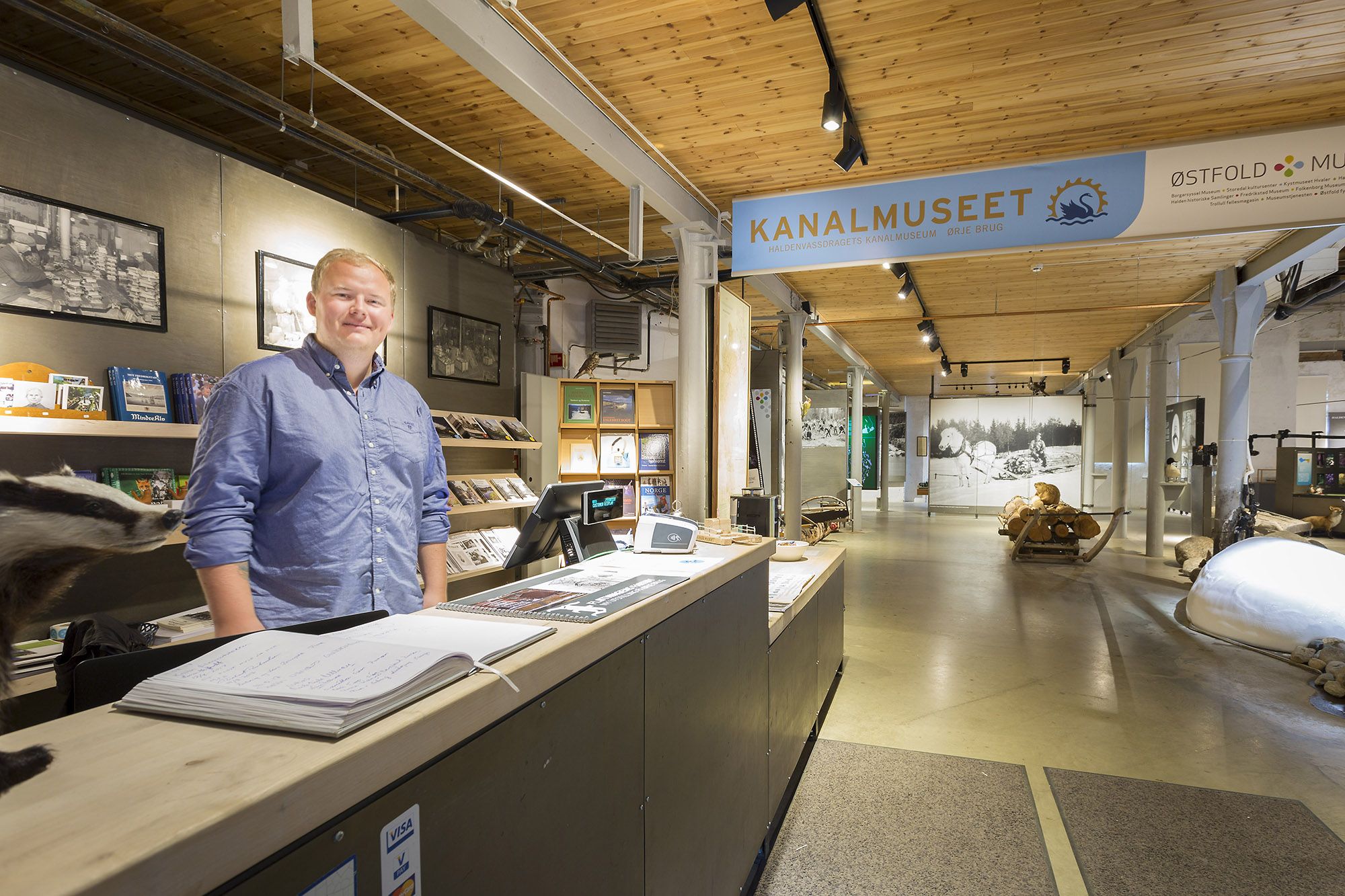 Bildet viser en mann som står bak resepsjonsdisken på Kanalmuseet. Han smiler mot kameraet. Rundt ham er brosjyrer, bøker og en datamaskin. I bakgrunnen kan man se museums utstillinger med bilder, båter og tømmerstokker.